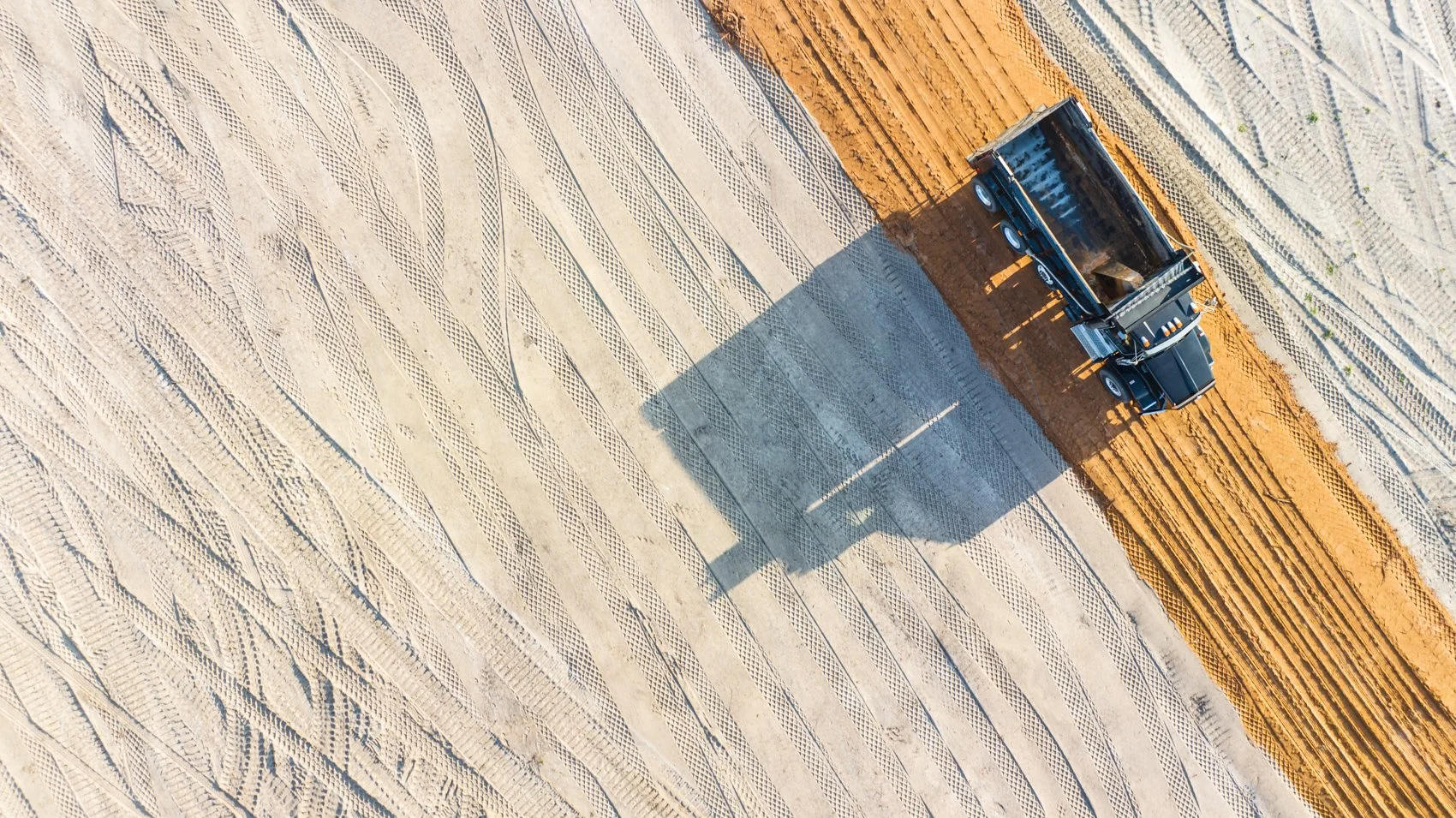 An aerial view of a construction area with a black dump truck parked on dirt, casting a large shadow on the sand and gravel surface.