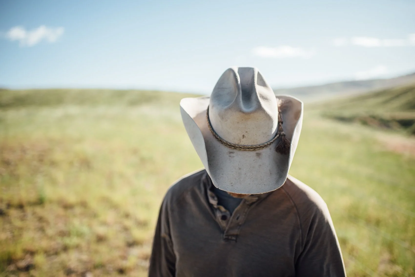 Person wearing a cowboy hat outdoors in a field with hills in the background.