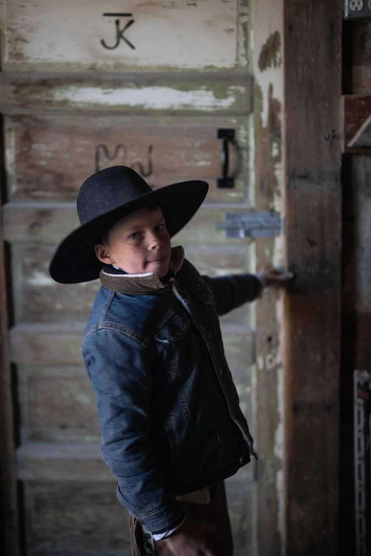 A young boy wearing a large black hat and denim jacket, standing in front of rustic wood pantry doors, looking at the camera.