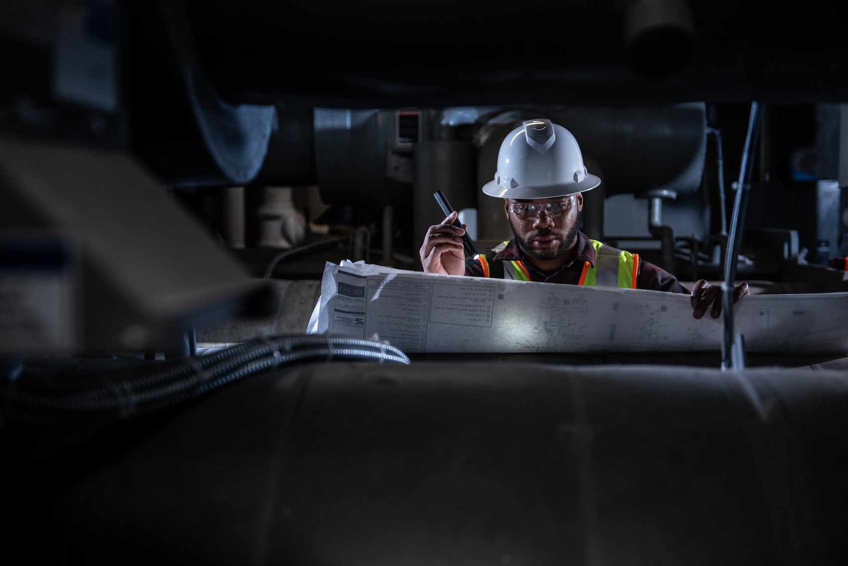 Construction worker in safety gear, hard hat, and safety glasses examining blueprints under low light.