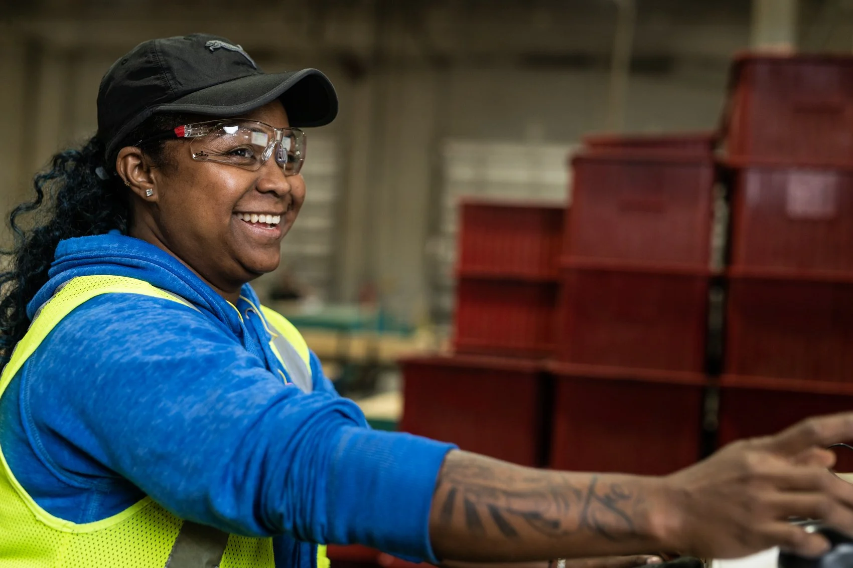 A woman with dark curly hair, wearing safety glasses, a black cap, a blue hoodie, and a yellow safety vest, smiling while working in a warehouse or industrial setting with red plastic storage bins in the background.