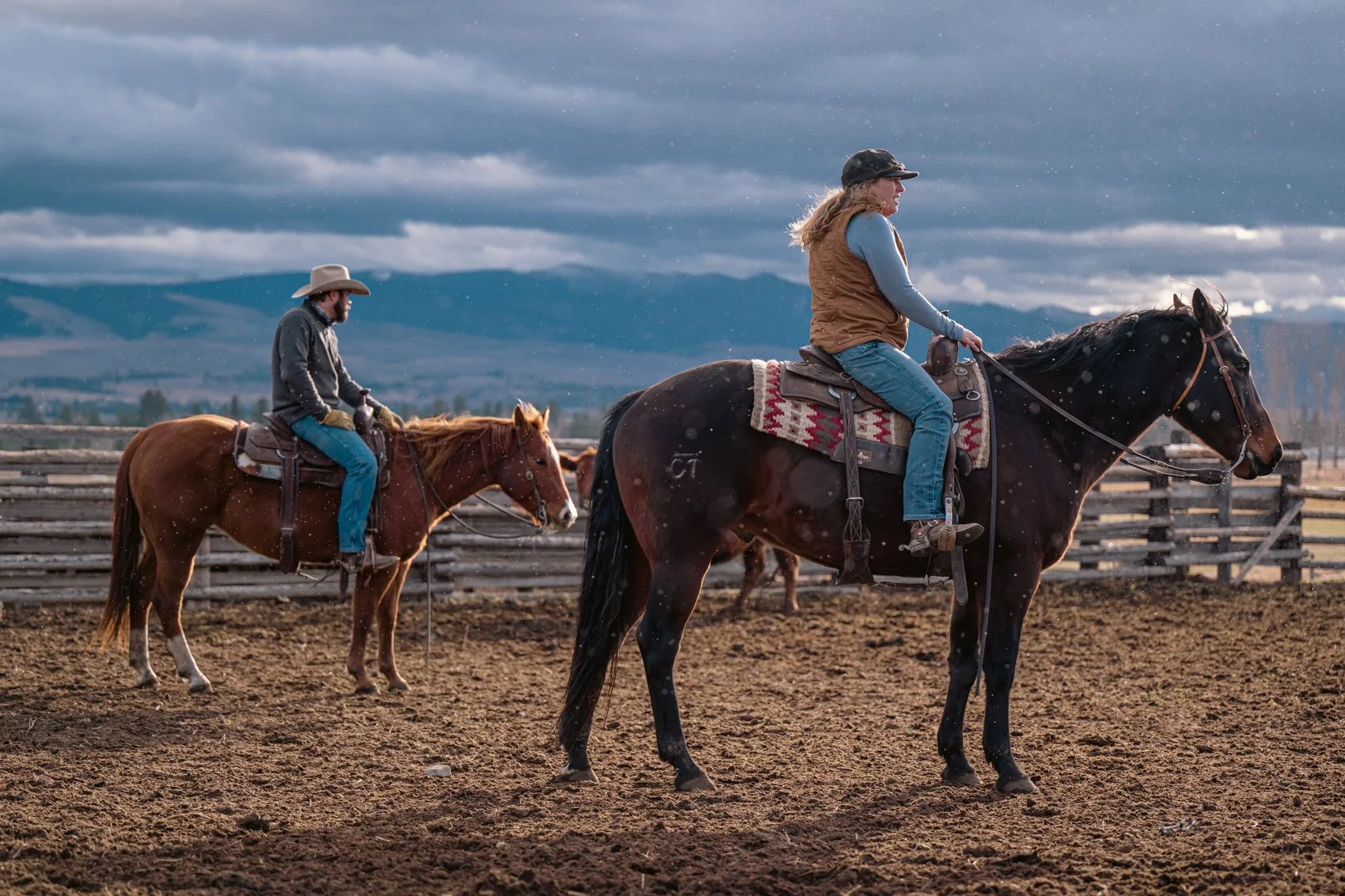 Two people riding horses in an outdoor riding arena with a wooden fence, under a cloudy sky.