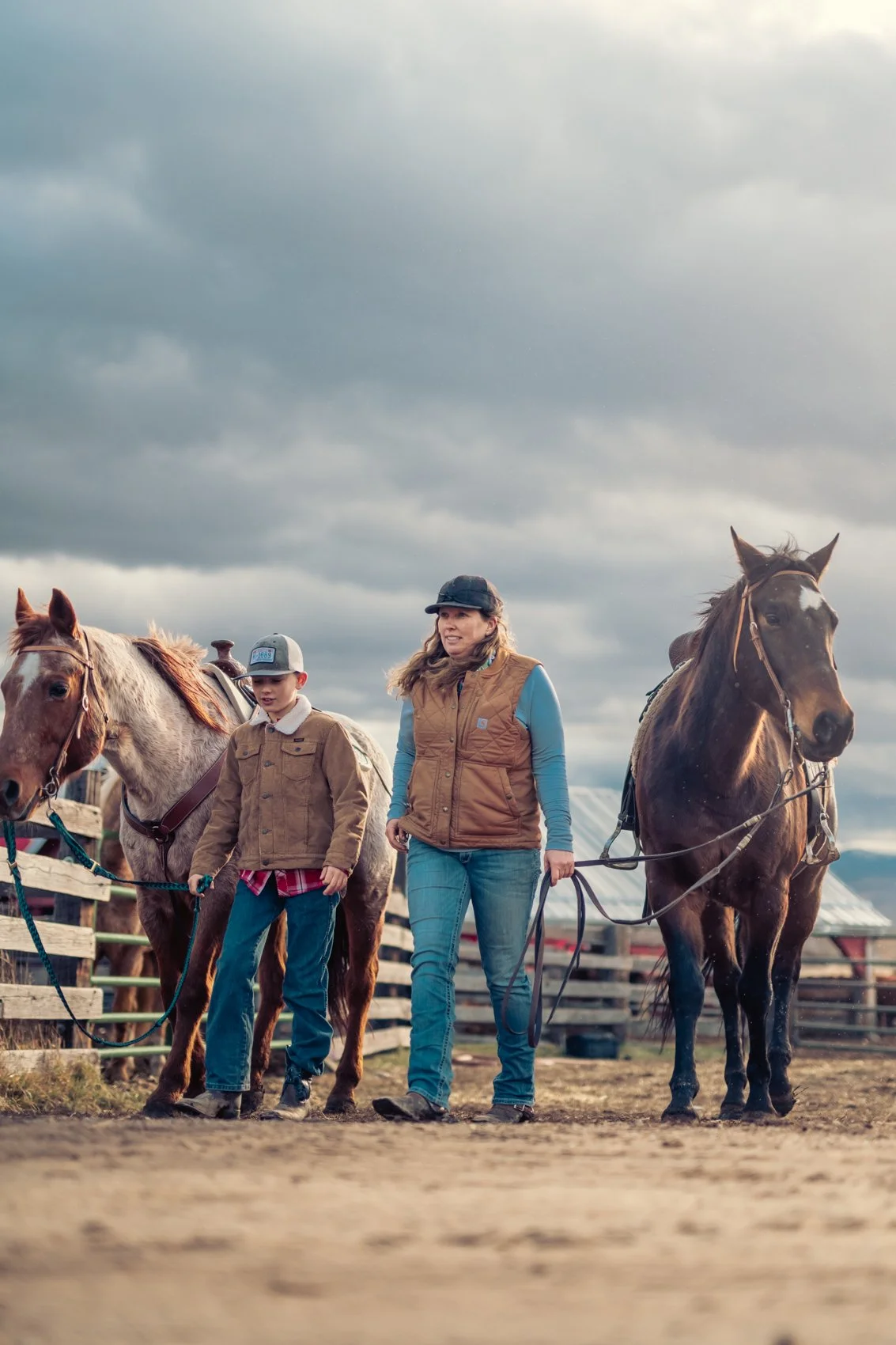 A woman and a boy walking with horses on a farm or ranch, under cloudy skies.