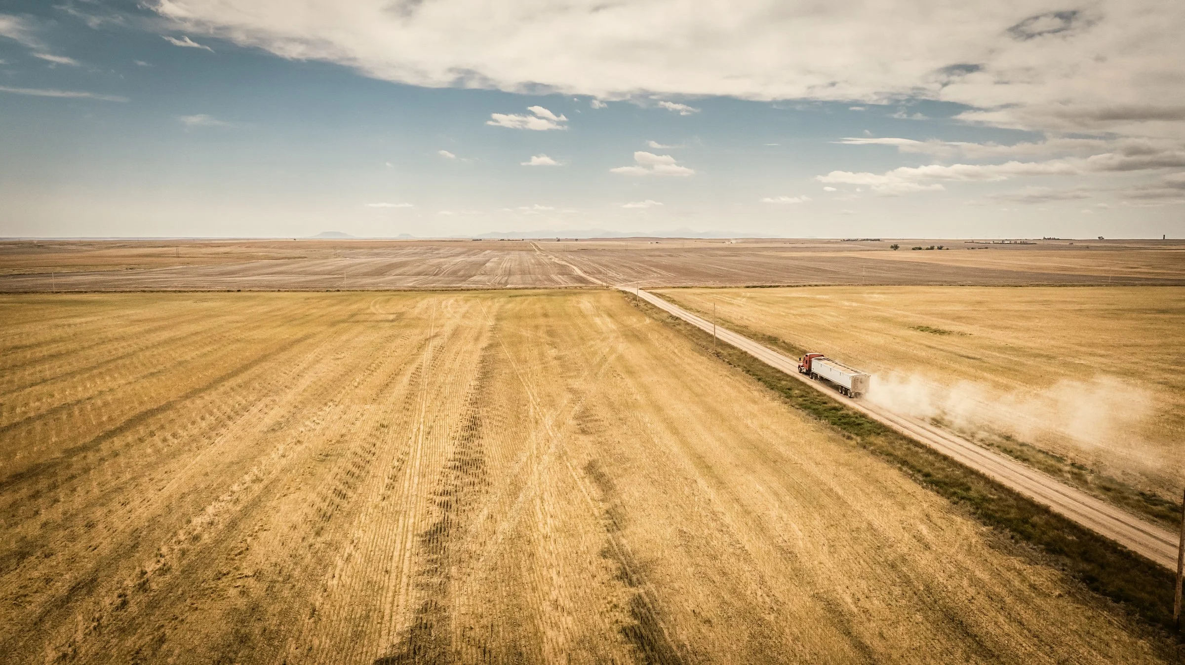 A long dirt road runs through vast golden fields on a clear day with scattered clouds, with a semi-truck traveling along the road and kicking up dust.