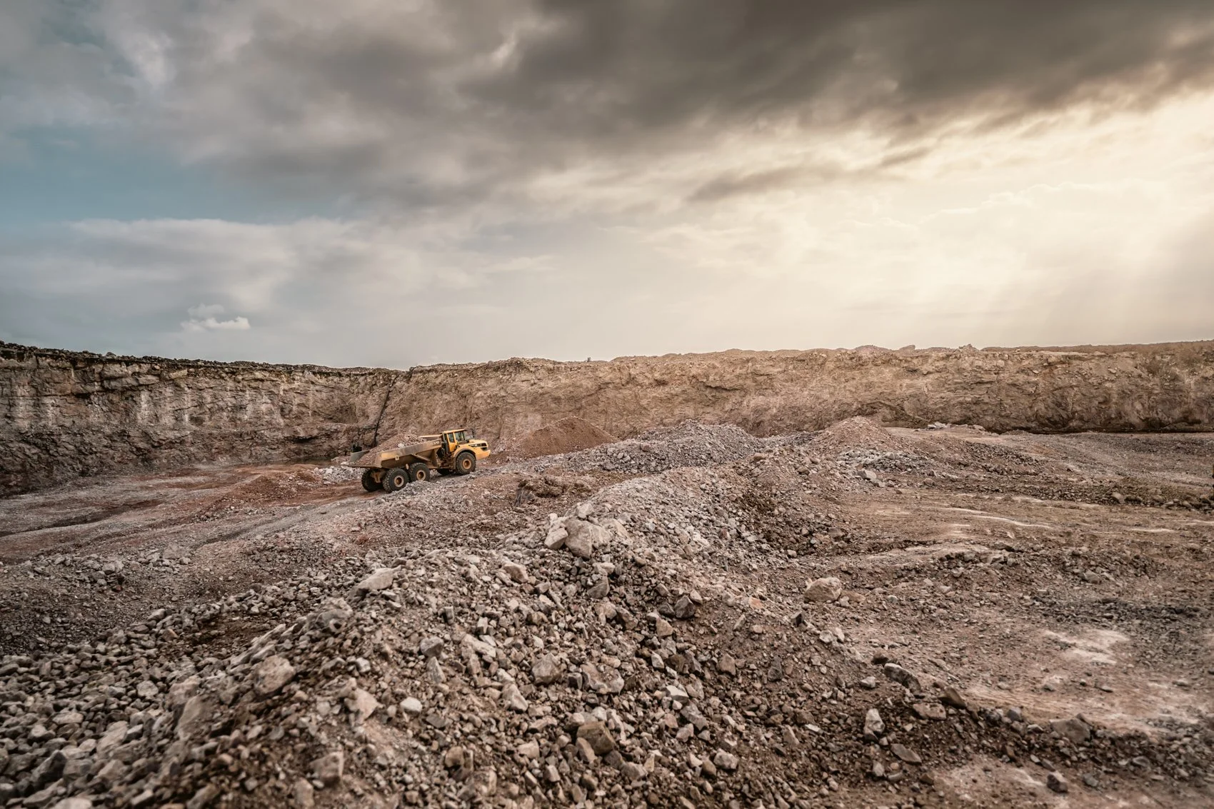 A large open-pit mining site with piles of rocks and a heavy-duty dump truck under a cloudy sky.