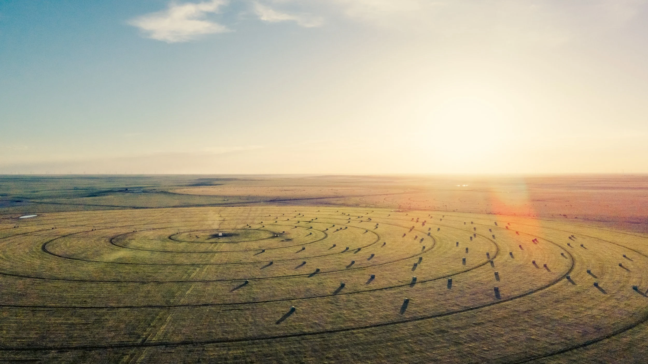 Aerial view of a large, flat field with circular crop growth patterns and a small building at the center, under a bright, setting sun.
