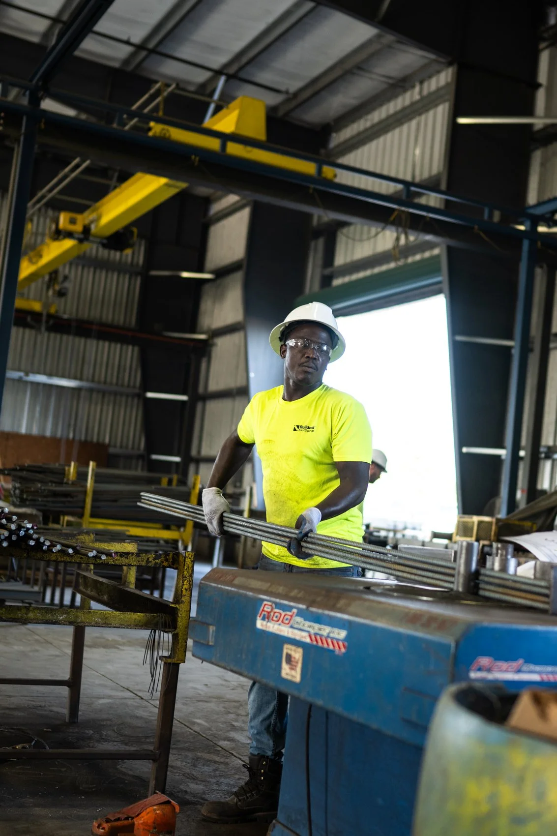 A worker wearing a yellow safety shirt, white hard hat, safety glasses, and gloves is inside an industrial workshop, handling metal rods. There are metal shelves and equipment around him.