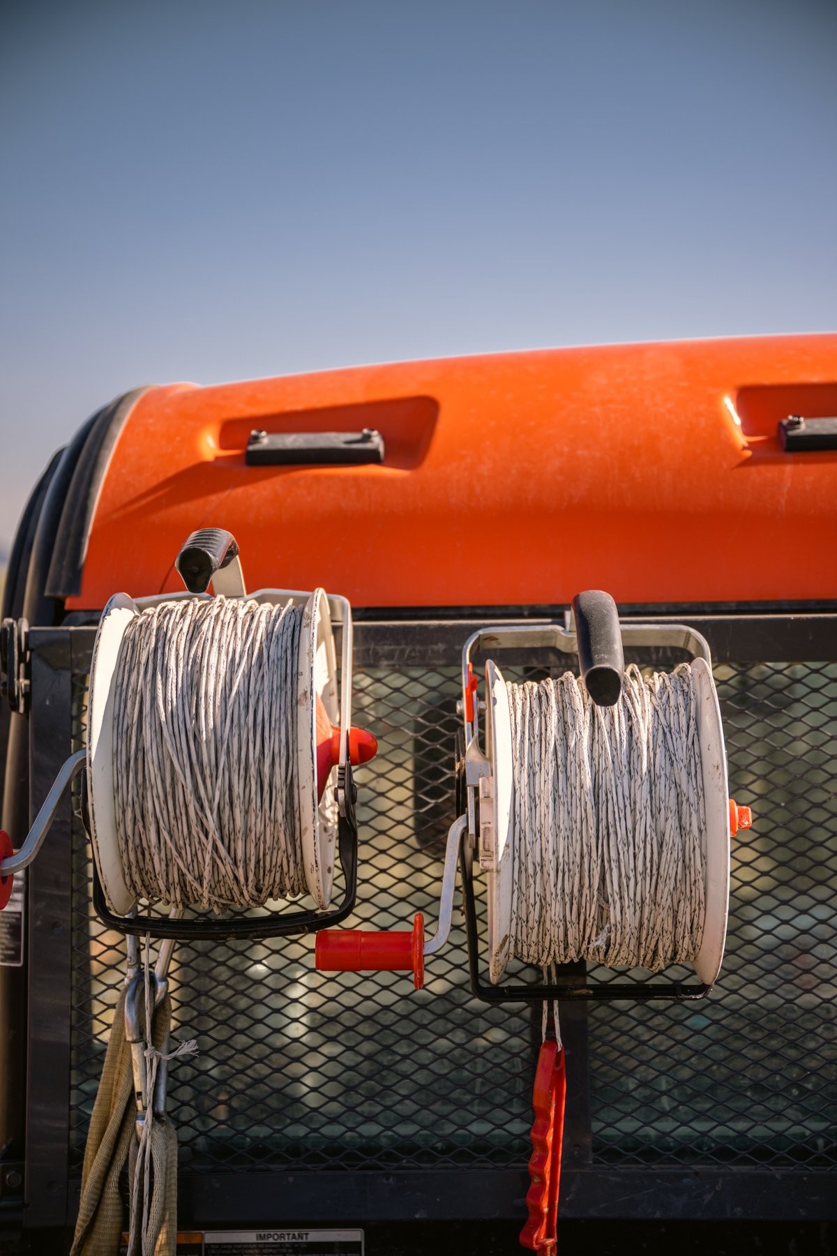 Close-up view of two hose reels with white coiled hose mounted on a metal grid surface, with an orange vehicle in the background and a clear blue sky.