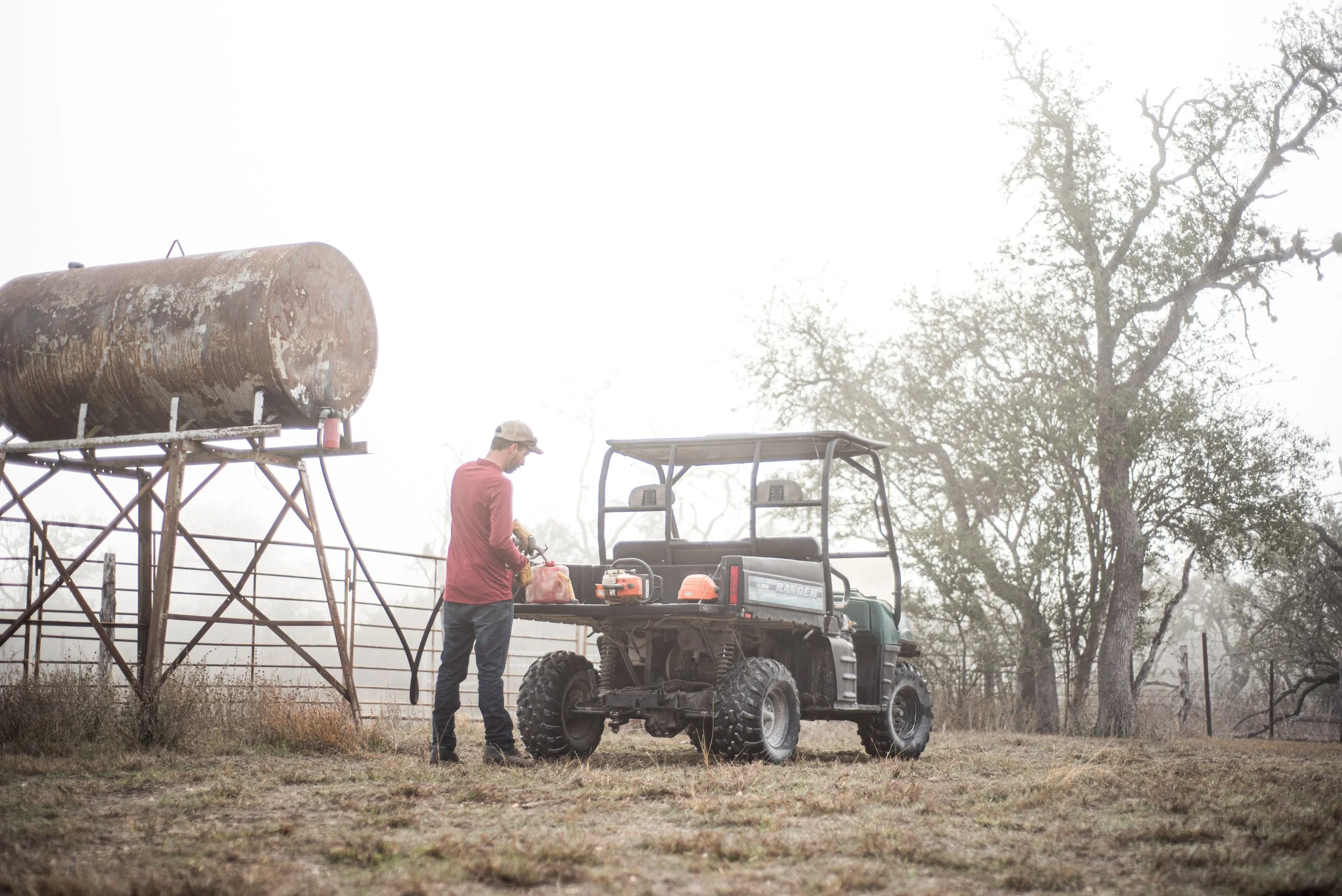 A man in a red shirt and beige cap filling a gas container at a small utility vehicle in a rural outdoor setting with trees and a rusted water tank.
