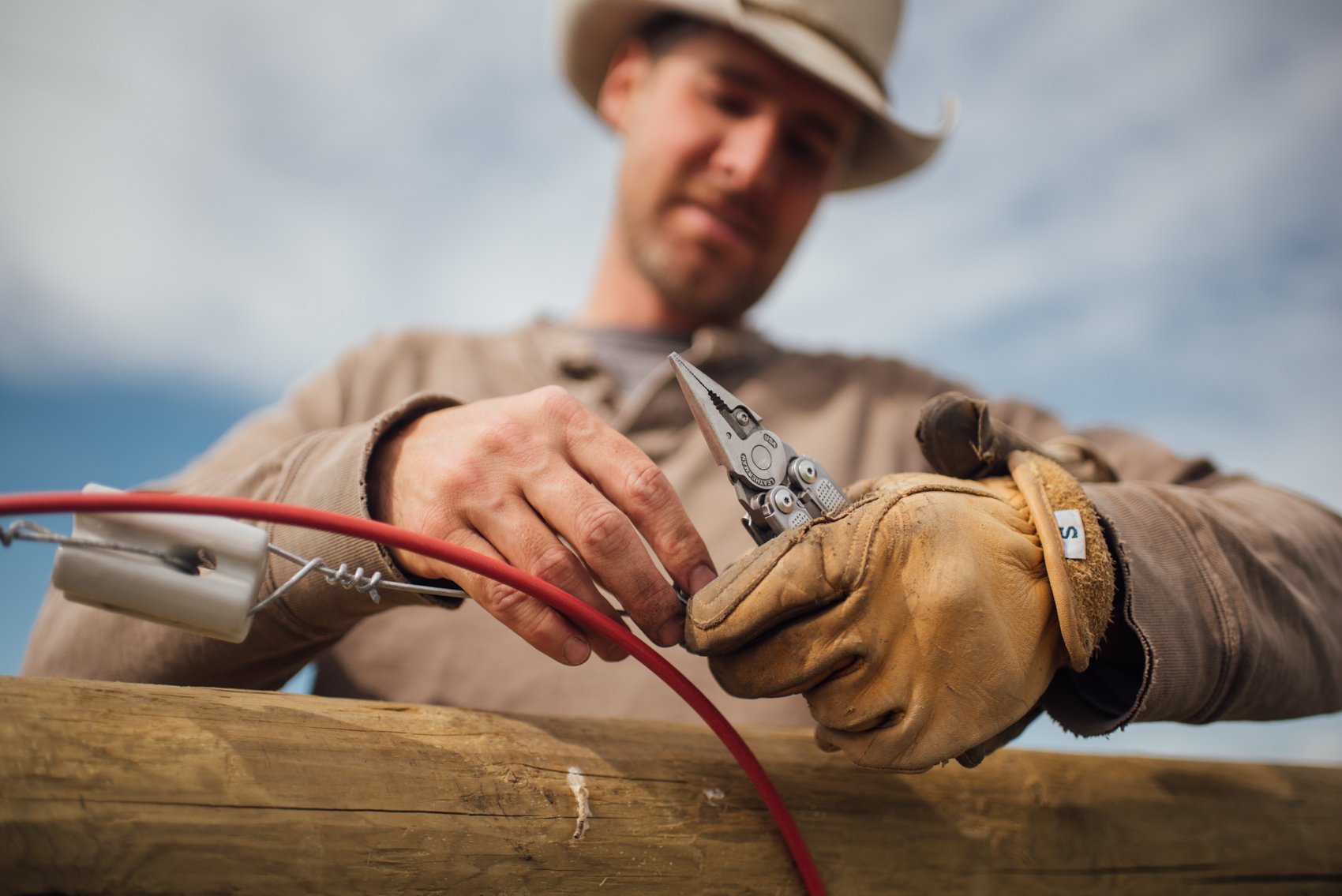 A man wearing a cowboy hat and gloves working with wire and pliers outdoors under a cloudy sky.