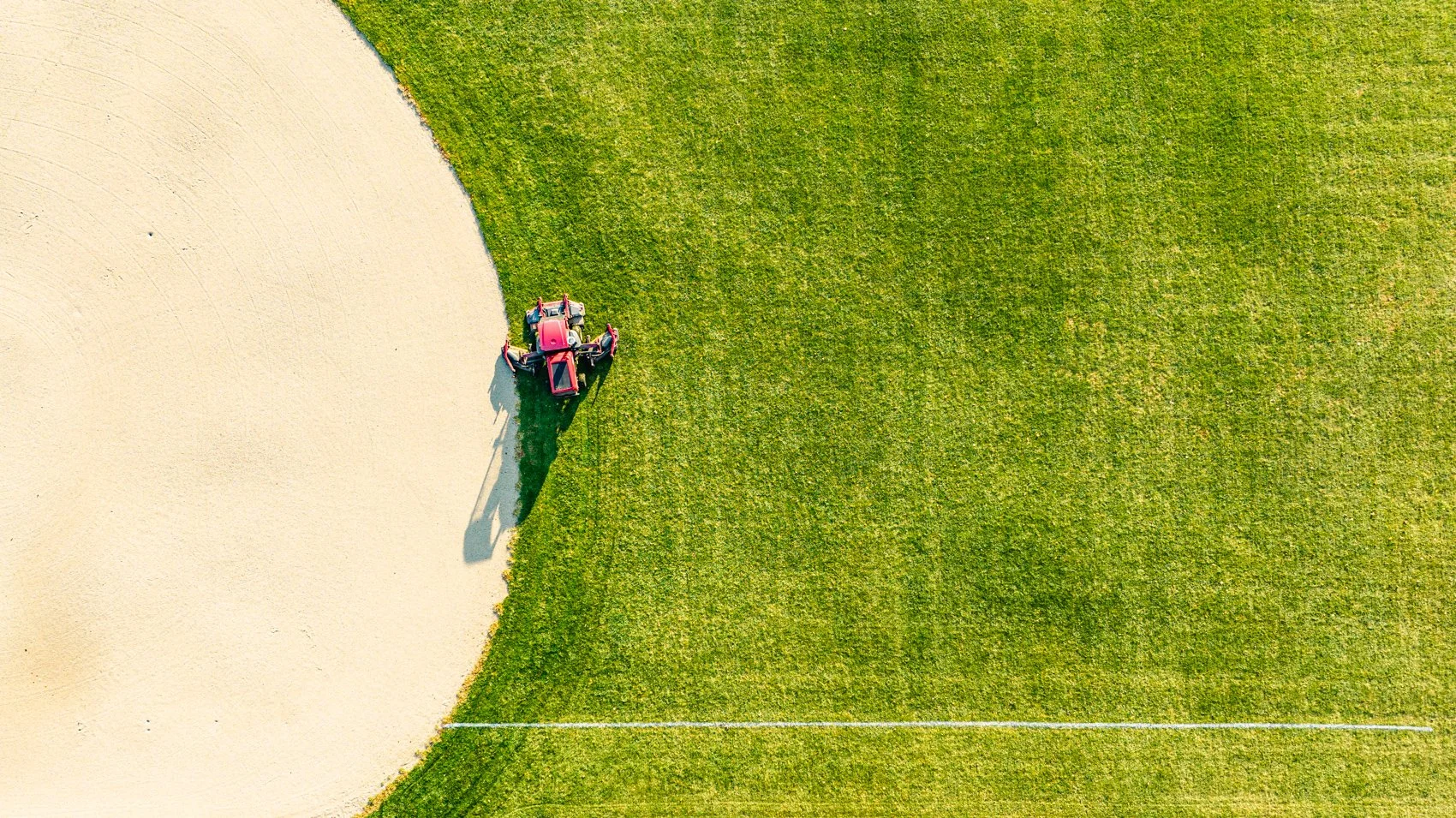 Aerial view of a red tractor working on a transition from green grass to a sandy area on a sports field.