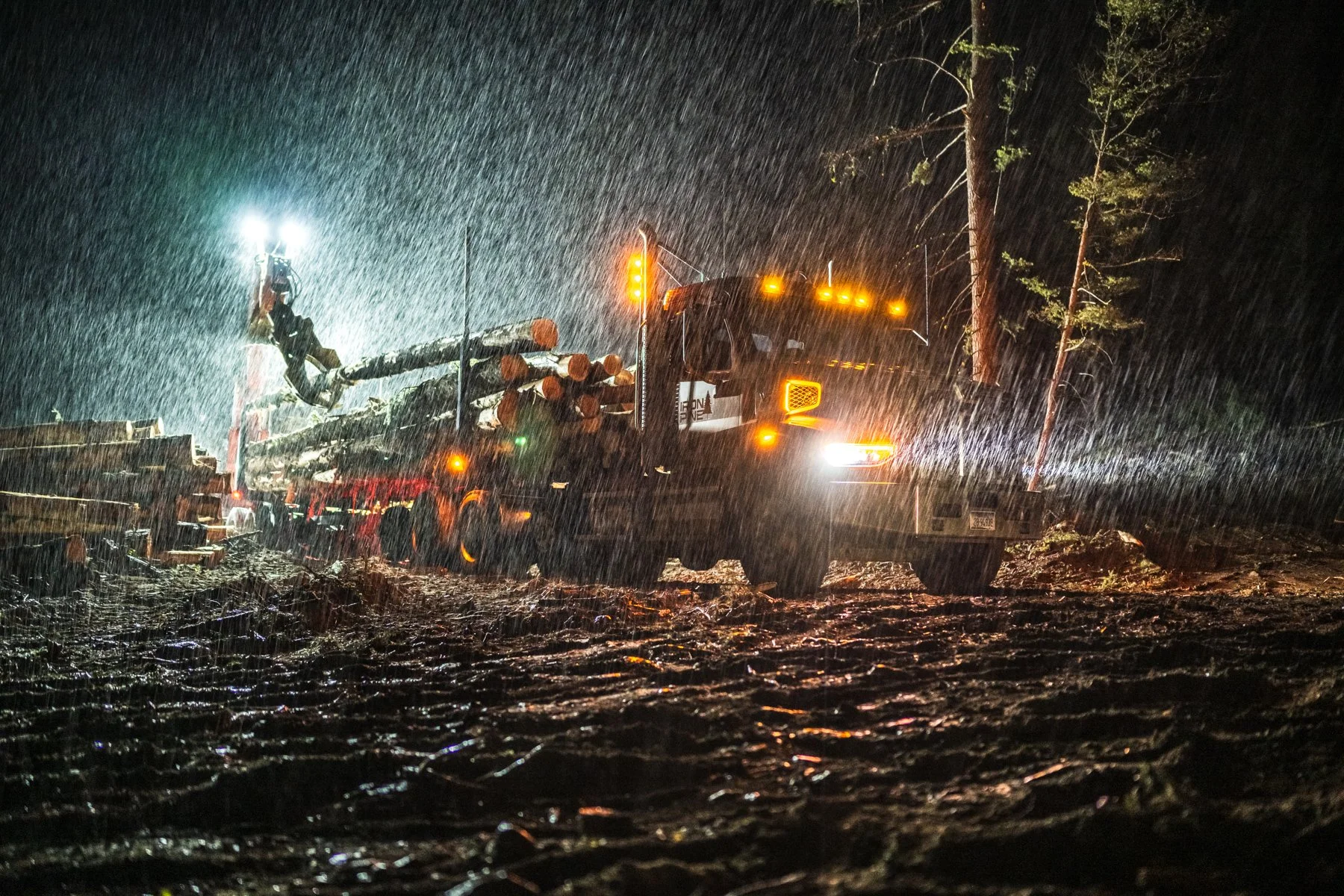 A logging truck is driving through a rainstorm at night, with logs on its flatbed and streetlights illuminating the scene.