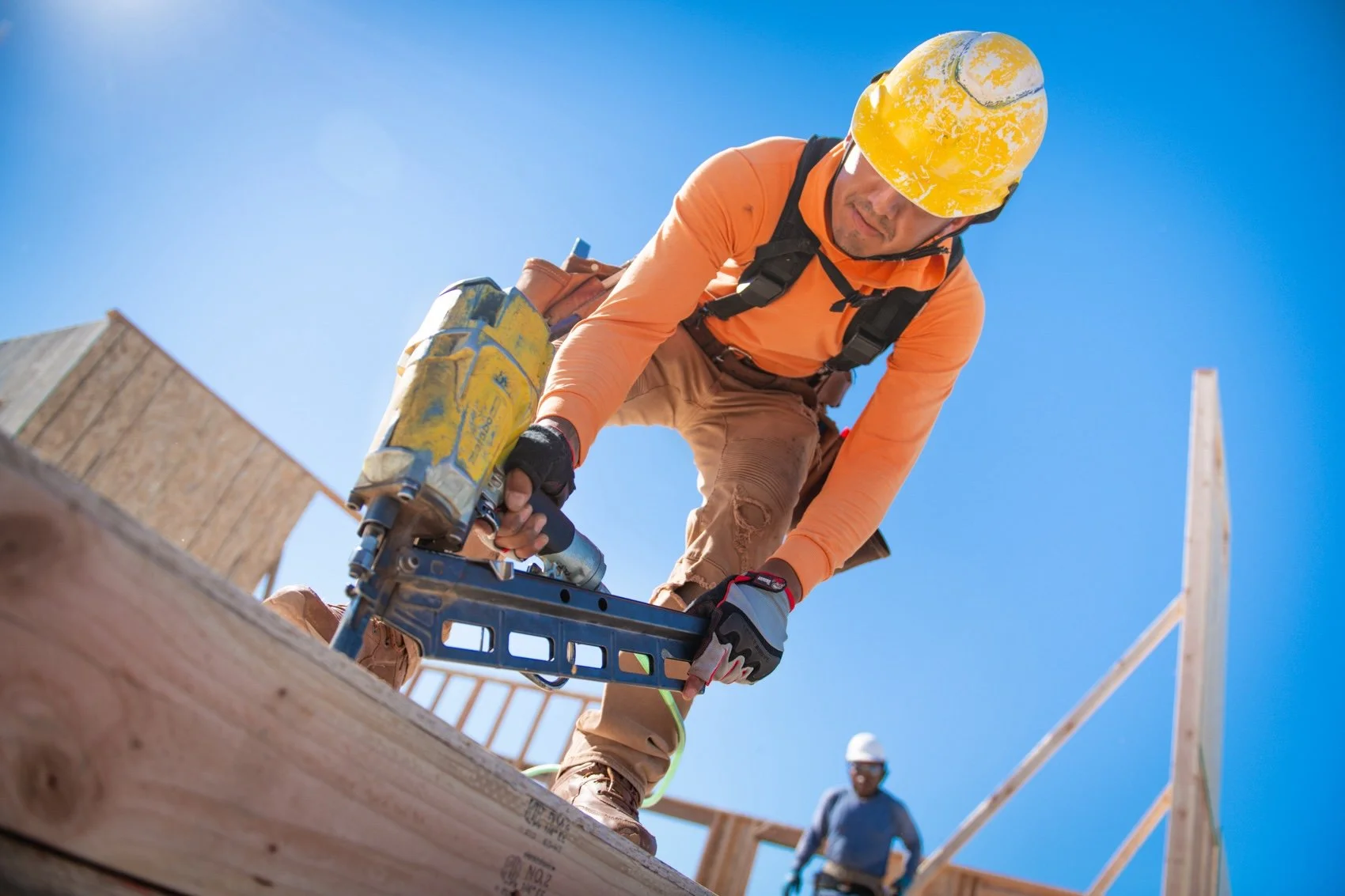 Construction worker wearing a yellow helmet and orange long-sleeve shirt using a pneumatic nailer on a wooden structure with clear blue sky background.