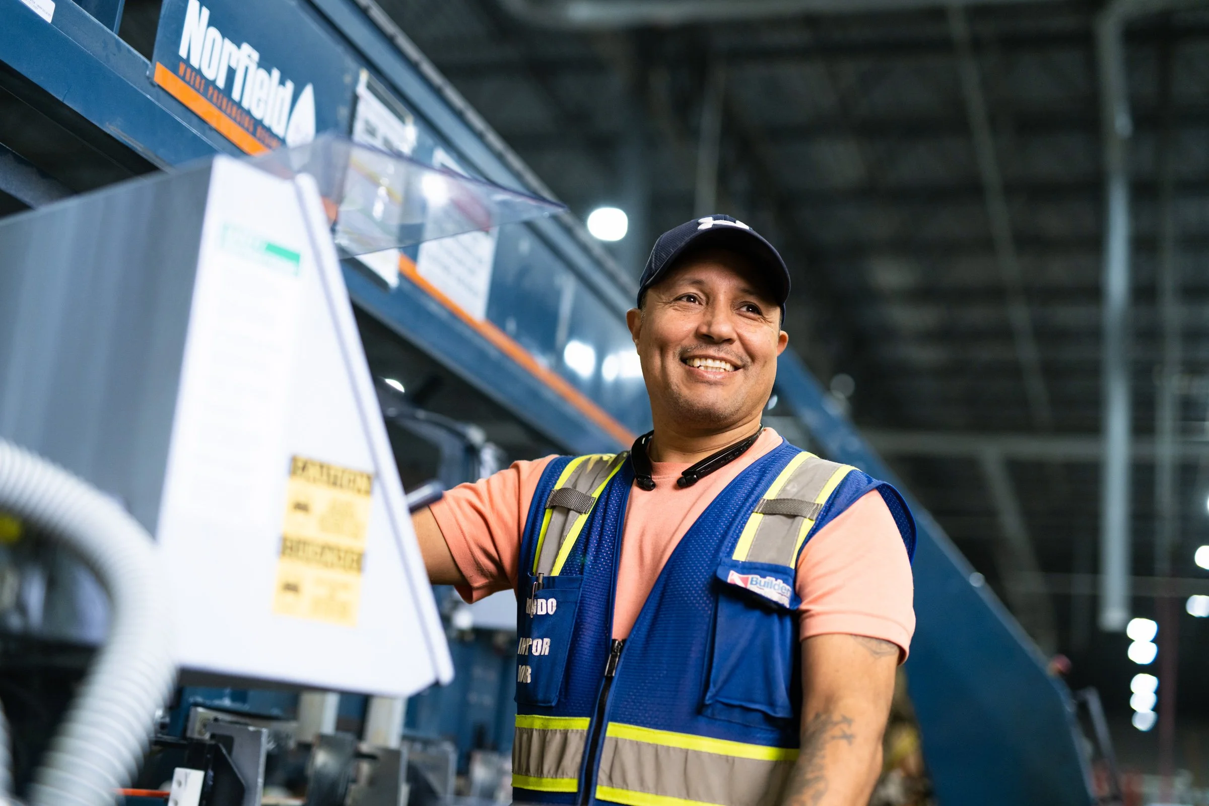 A smiling man in a construction vest and cap at a warehouse or industrial facility.