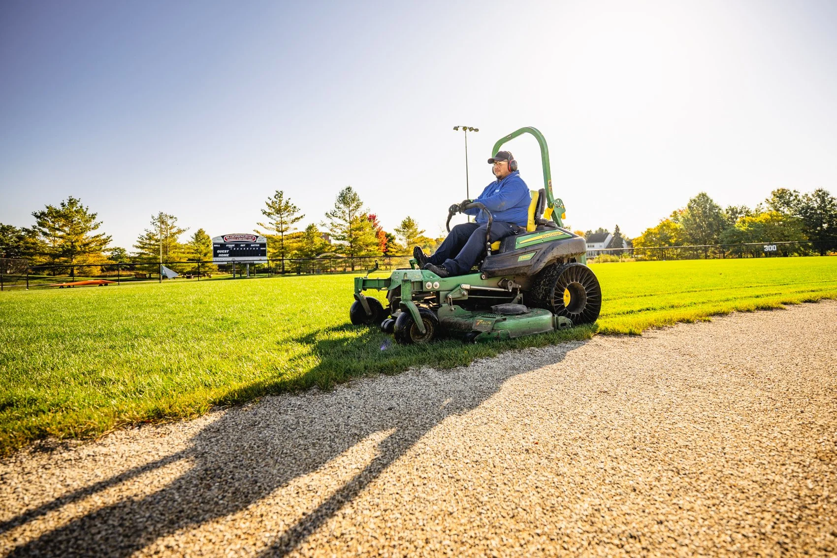 Man operating a grass mower on a sunny field with trees and scoreboard in the background.