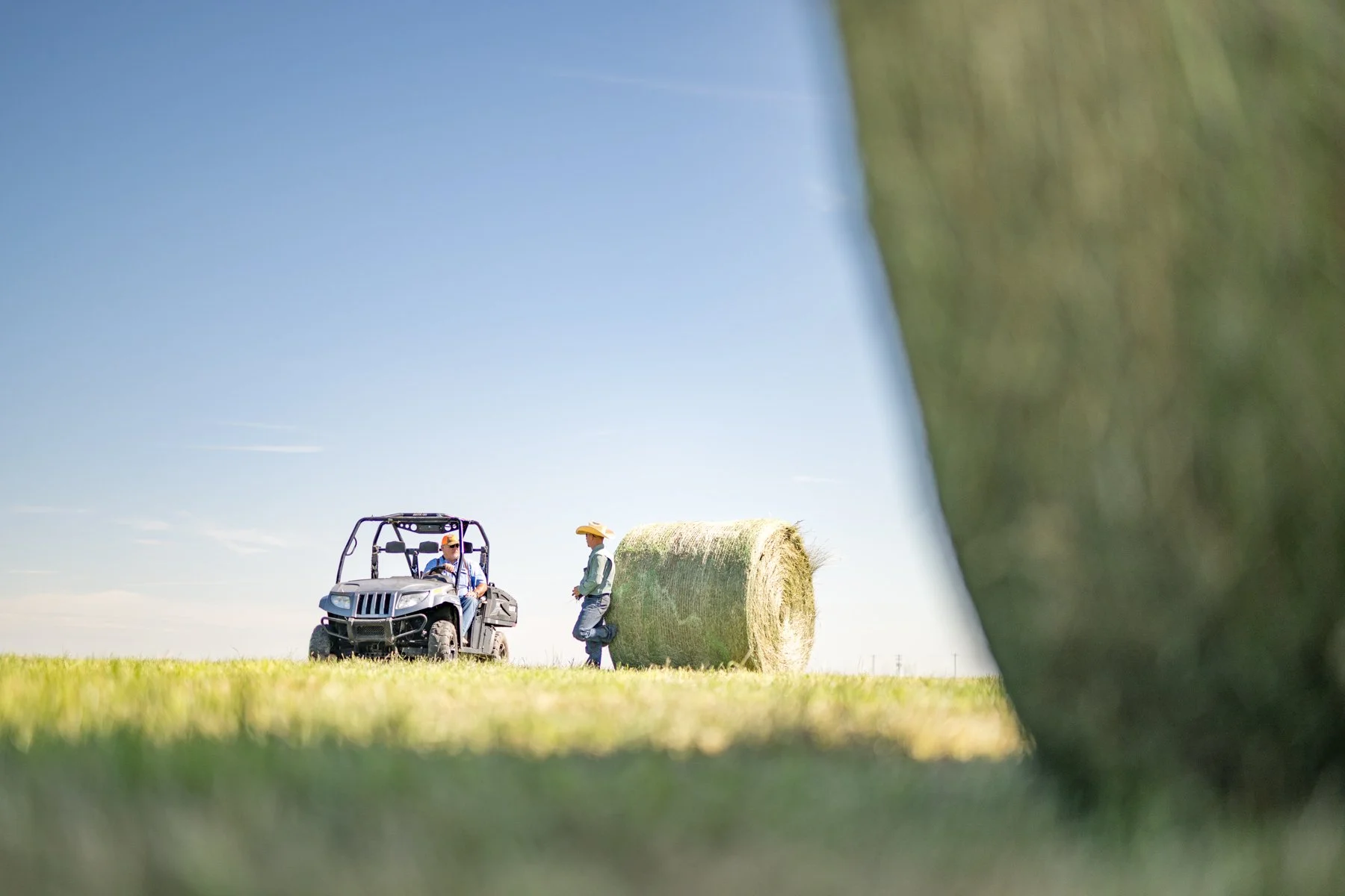 People on a tractor talking to a man with a cowboy hat near a hay bale in a grassy field under a blue sky.