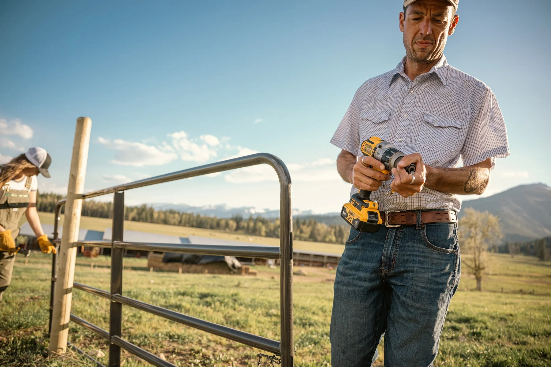 A man in jeans and a checkered shirt using a drill outdoors, with another person working in the background on a farm with a fence and open field, mountains and blue sky in the distance.