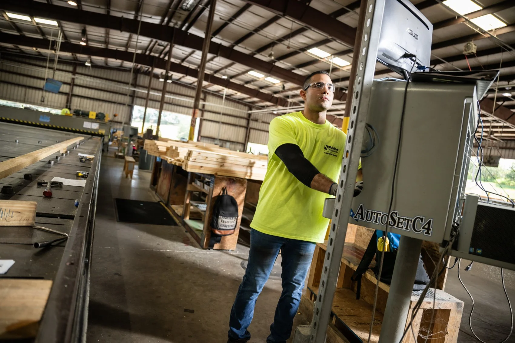 A worker in a warehouse wearing safety glasses and a yellow T-shirt operating a computer in front of industrial equipment, with stacks of wood around.