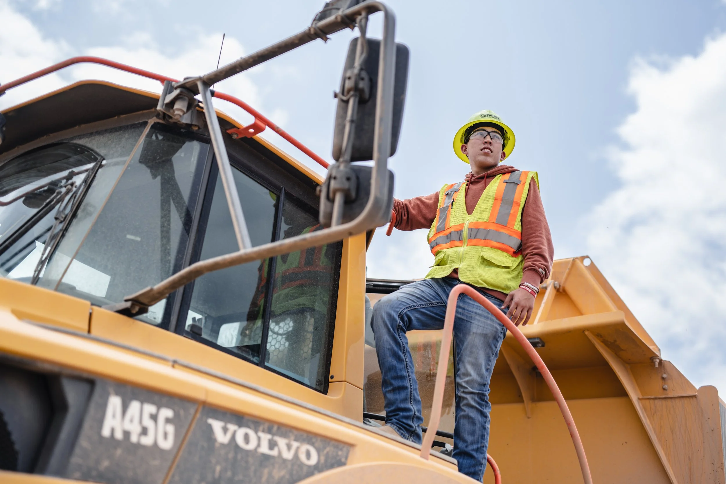 A young construction worker wearing a yellow safety helmet, safety vest, glasses, brown hoodie, and jeans sits on the side step of a yellow Volvo A45G articulated dump truck on a construction site.