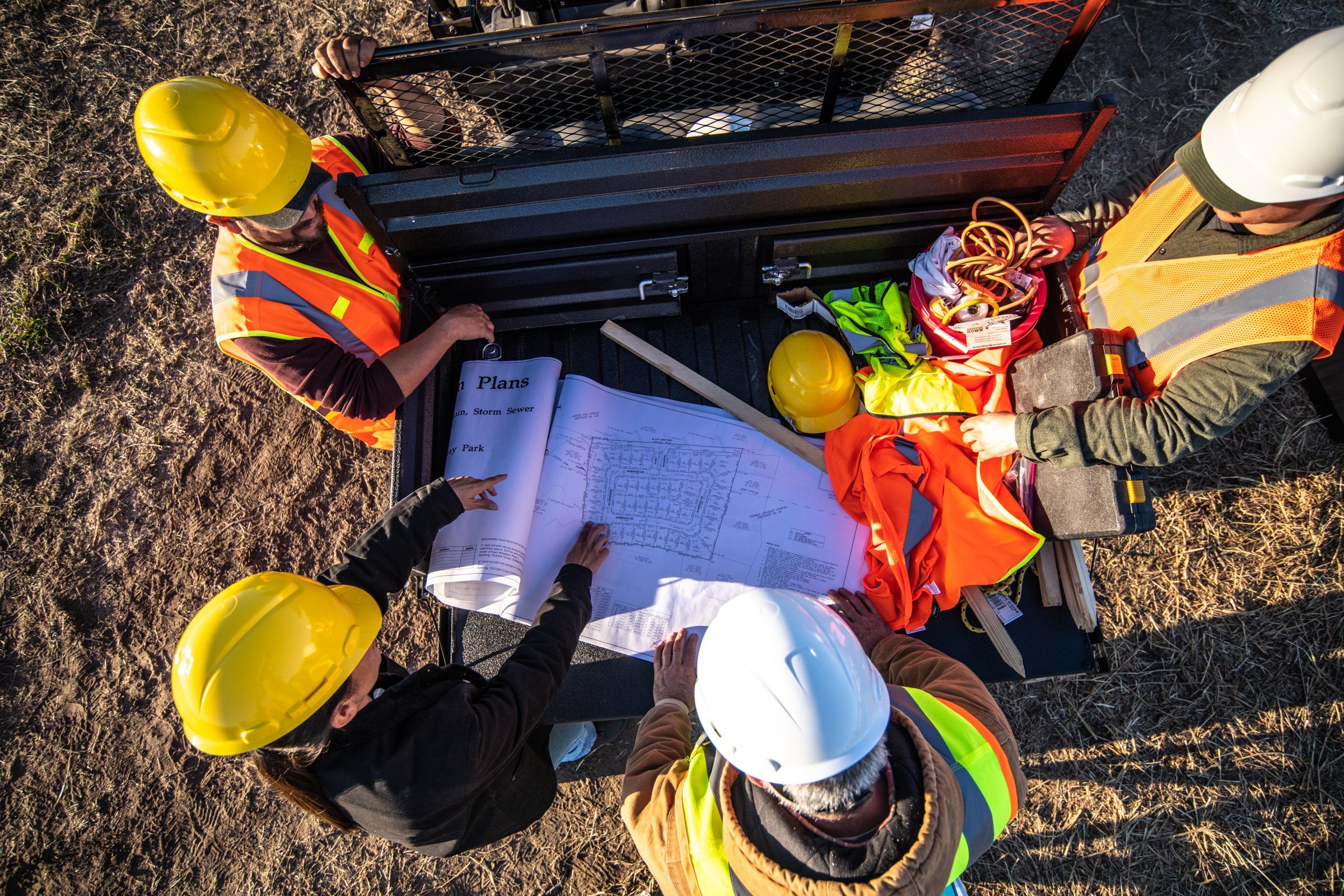 Four construction workers wearing safety helmets and vests reviewing blueprints and construction plans outdoors.