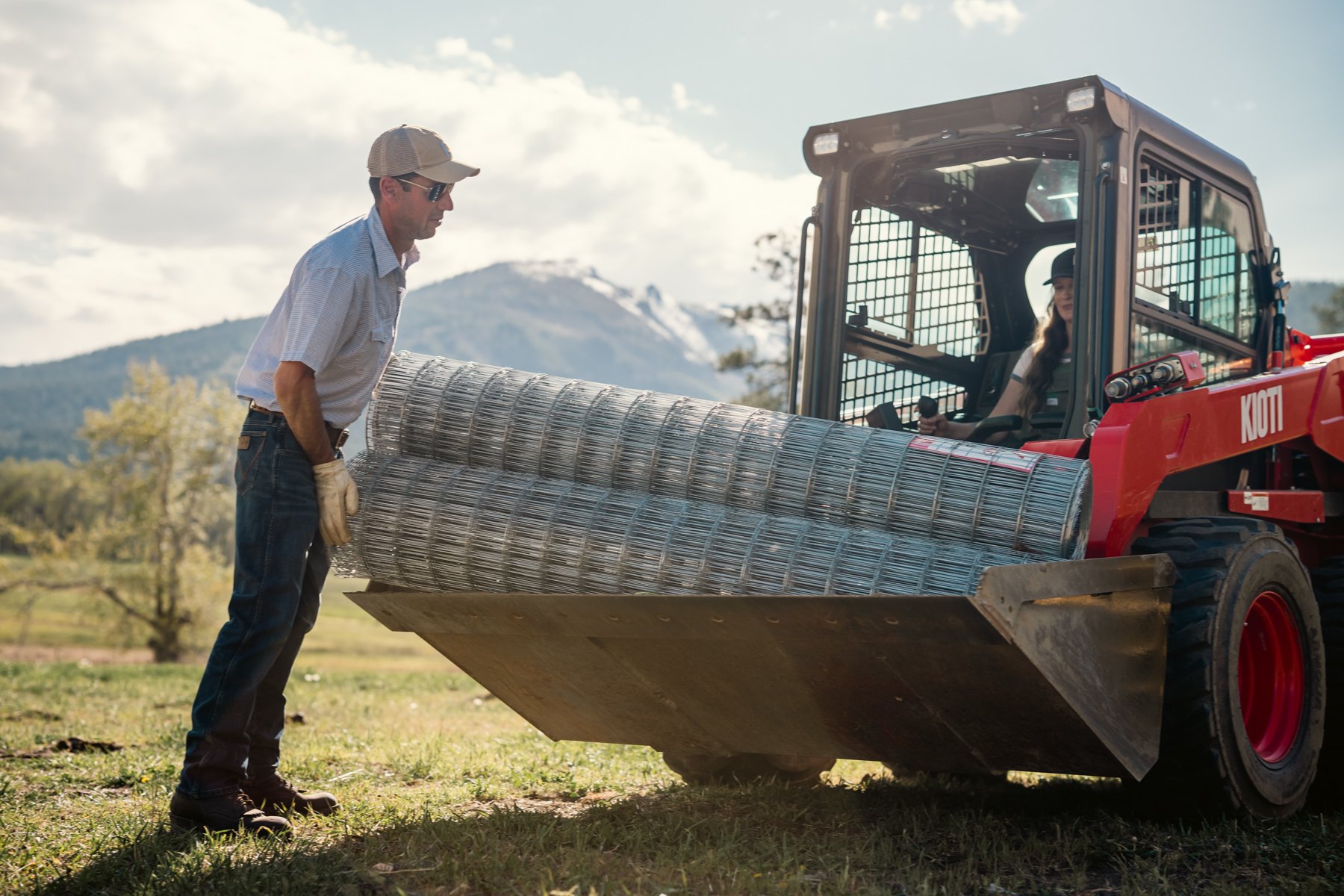 A man in a beige hat and sunglasses is helping a woman in the driver’s seat of a red forklift to place a large coiled metal wire roll onto the forklift.