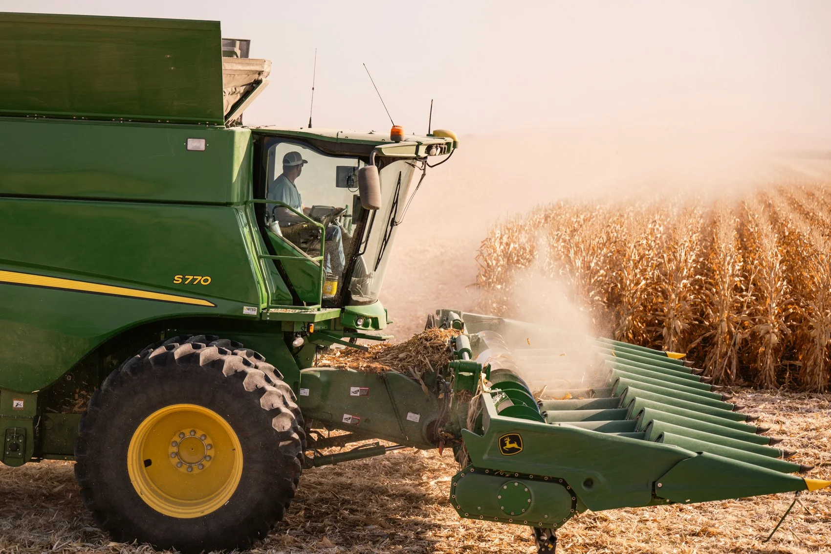 A green John Deere S770 combine harvester working in a cornfield, harvesting crops, with dust rising in the field during sunset.