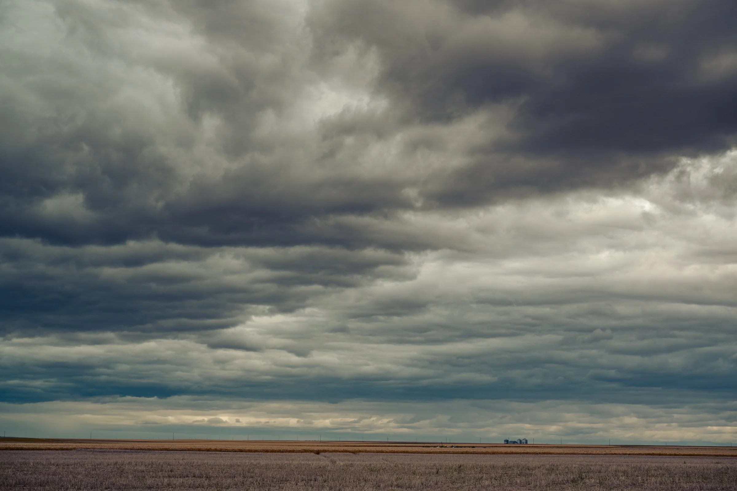 A wide-open plain with a dark, cloudy sky overhead and a distant farm building or silos on the horizon.