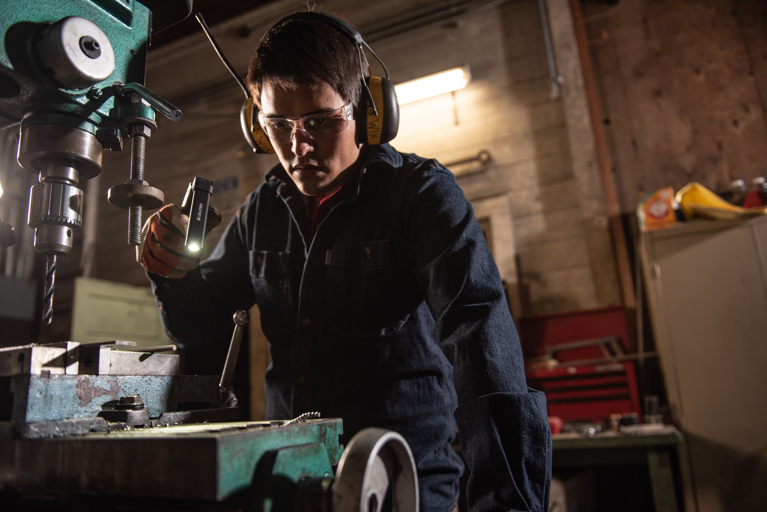 A young man in safety glasses and ear protection working on a drill press in a workshop.