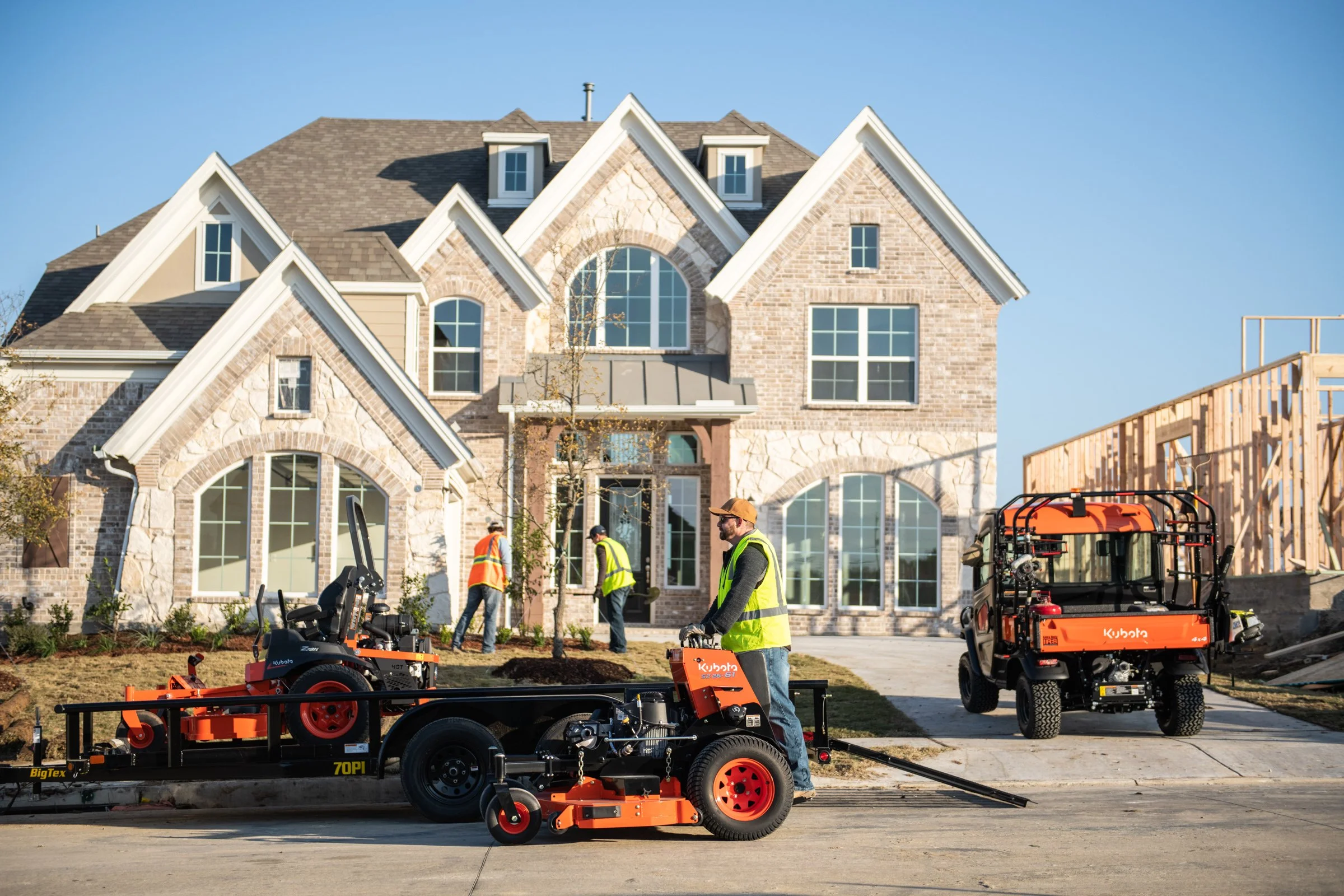 Construction workers in high-visibility vests working outside a large brick house during daytime, with construction equipment visible in the foreground.