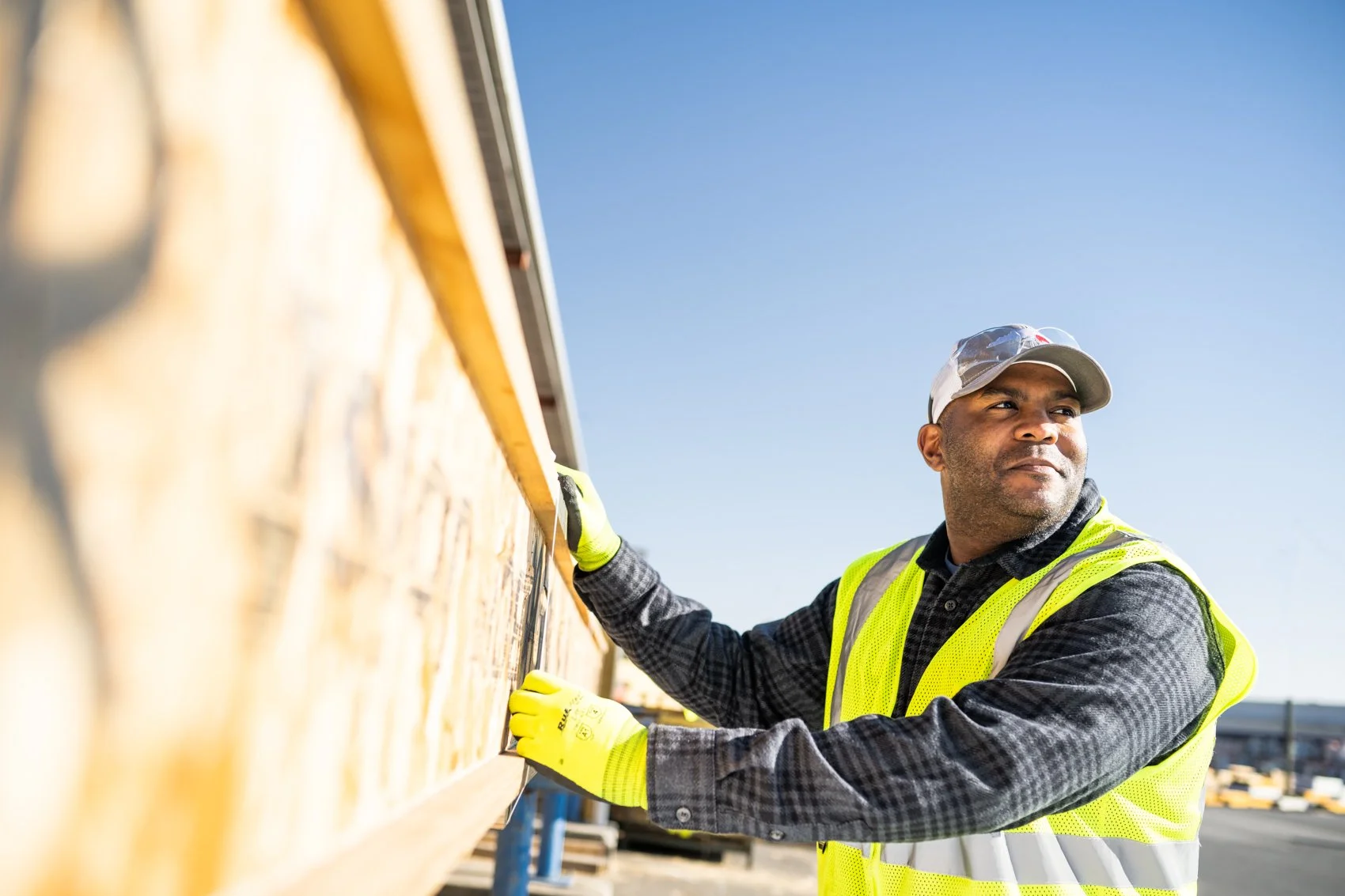 A construction worker wearing a yellow safety vest and gloves inspecting a wooden structure outdoors under a clear blue sky.