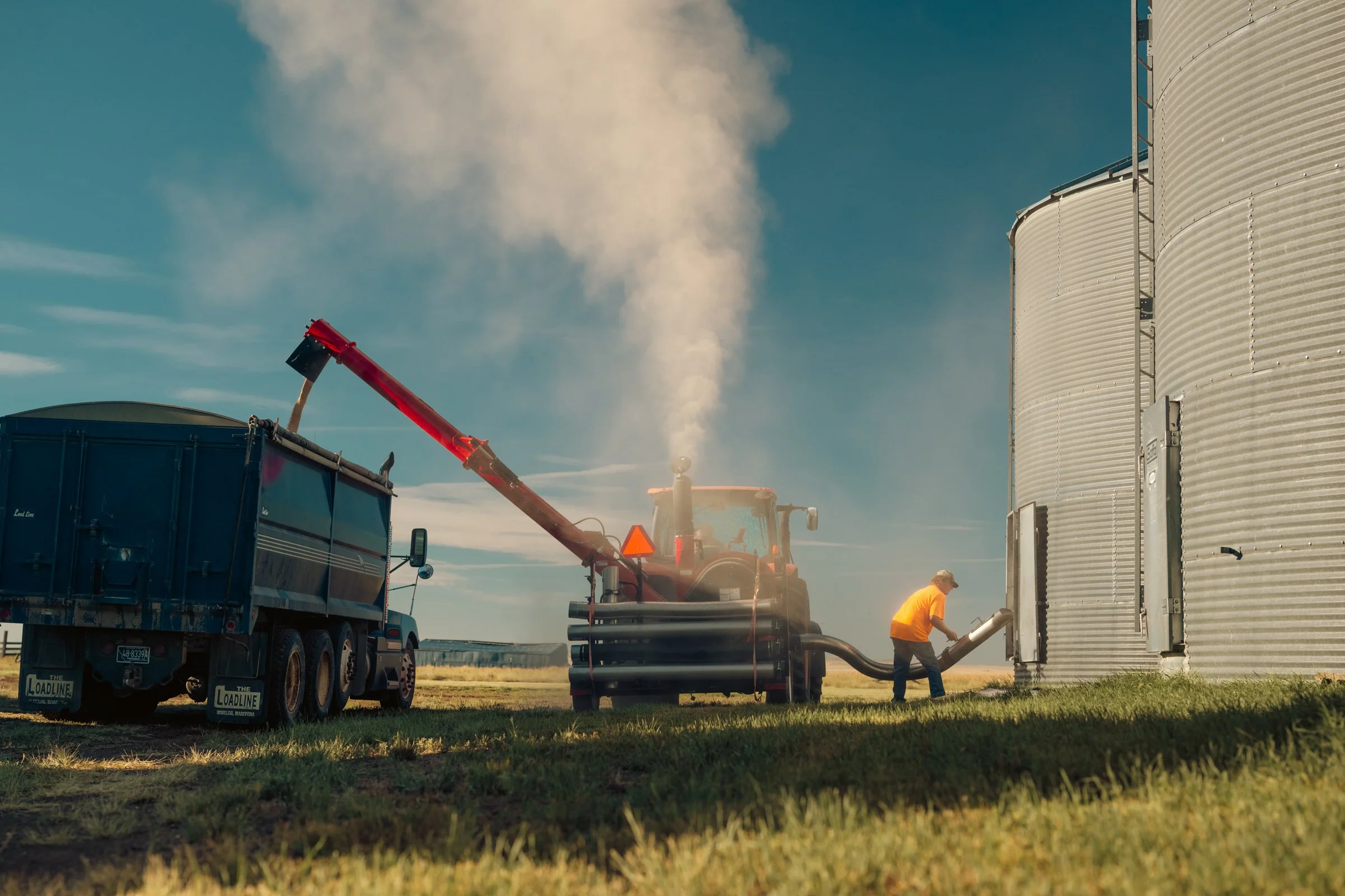 A person is pouring grain from a truck into metal silos on a farm under a clear blue sky.