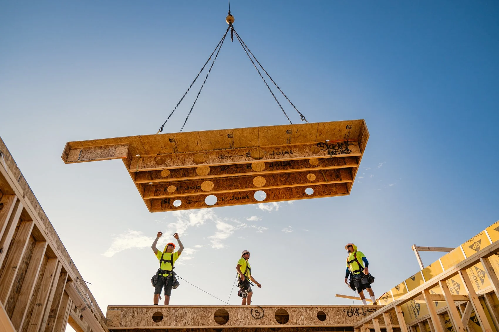 Construction workers in safety vests and helmets installing a large wooden beam on a building under construction, with clear blue sky in the background.
