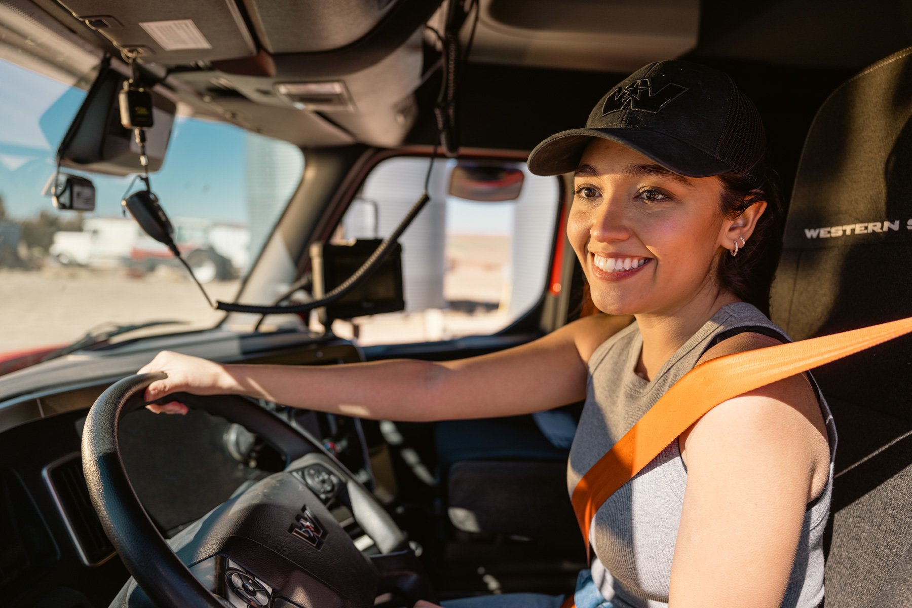Young woman smiling while driving a truck, wearing a gray sleeveless top and a black cap, with a seatbelt across her shoulder.