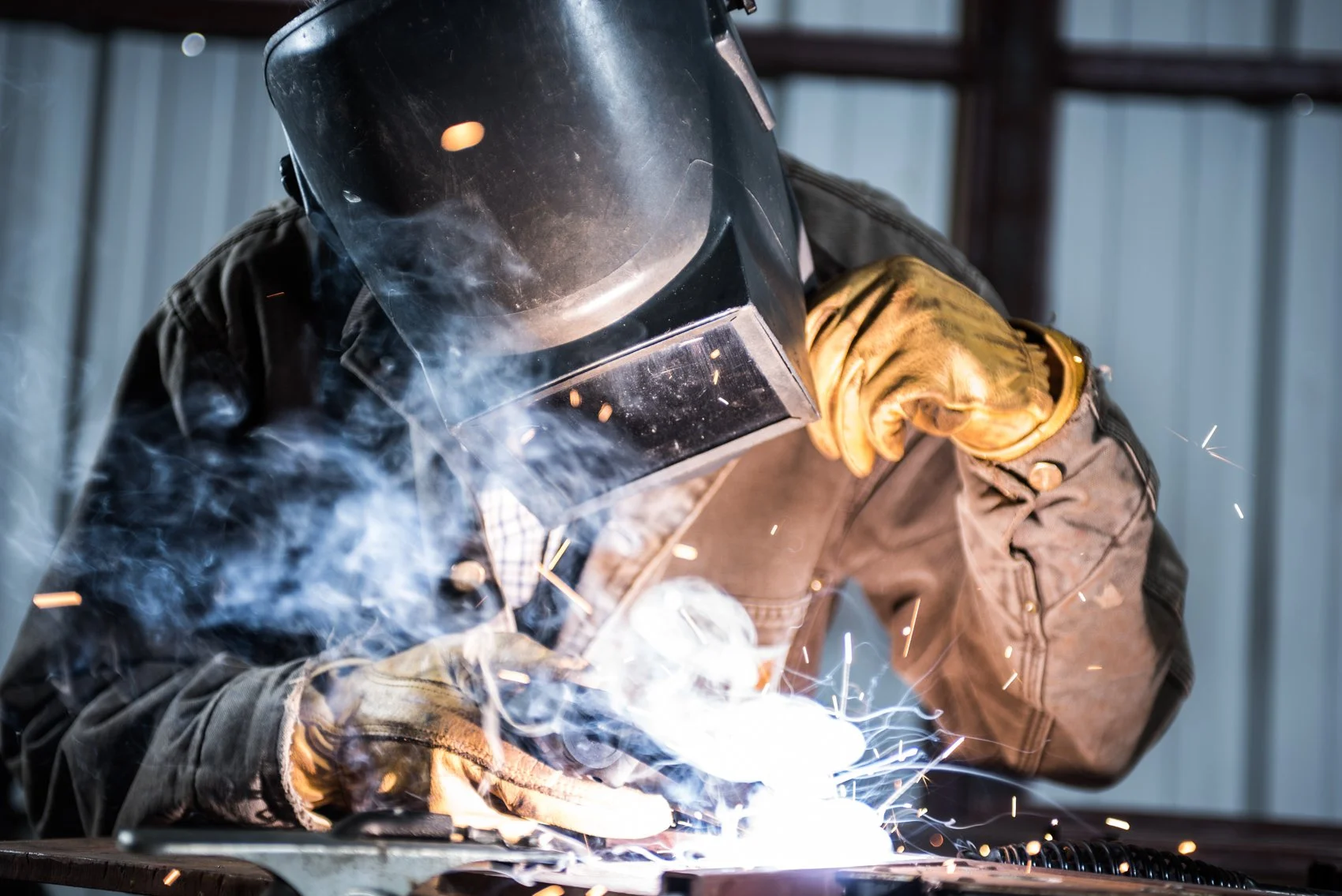 A person welding metal with a welding mask, gloves, and a brown jacket, sparks and smoke visible around.