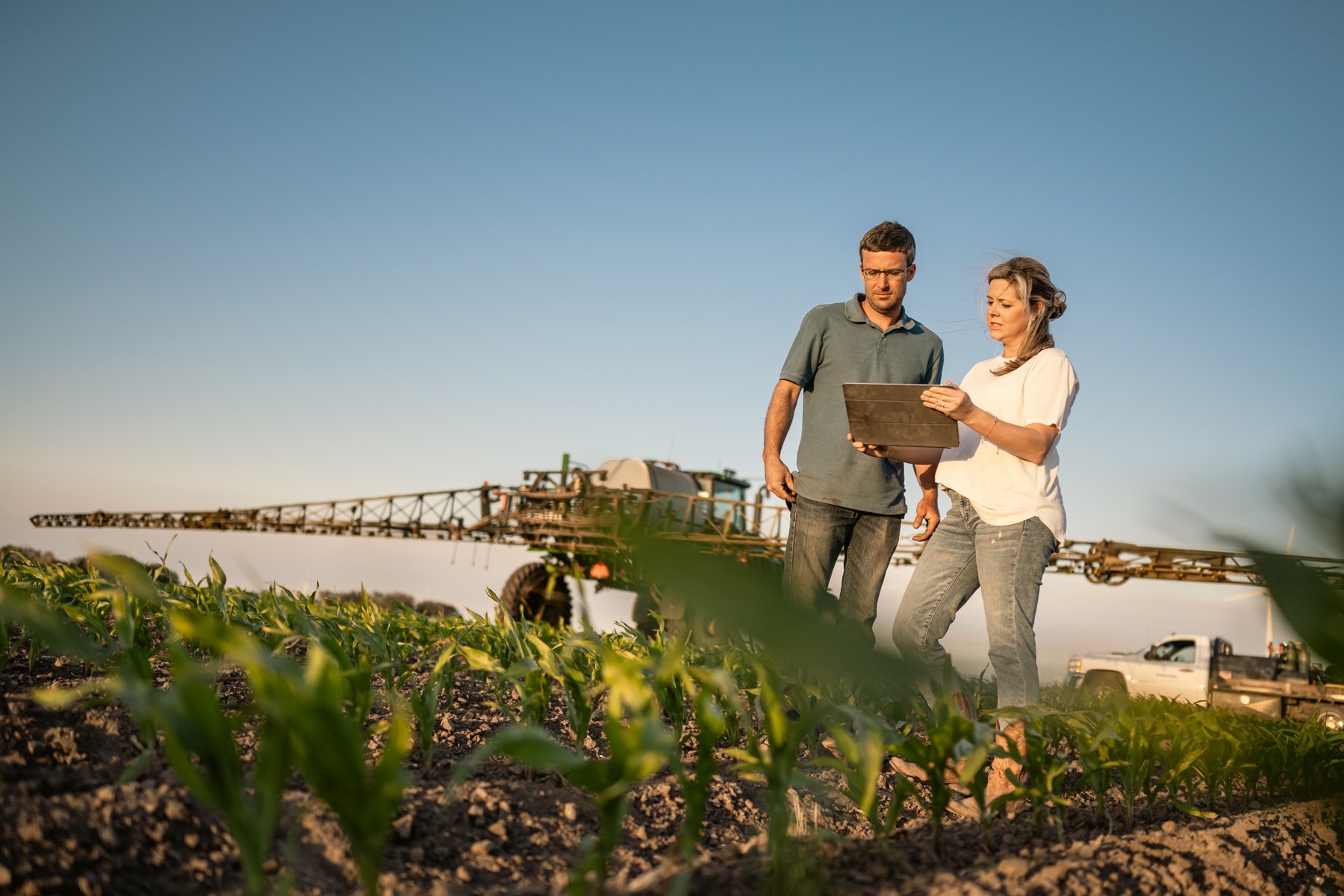Two farmers standing in a field, reviewing a tablet, with farm equipment and a pickup truck in the background under a clear sky.