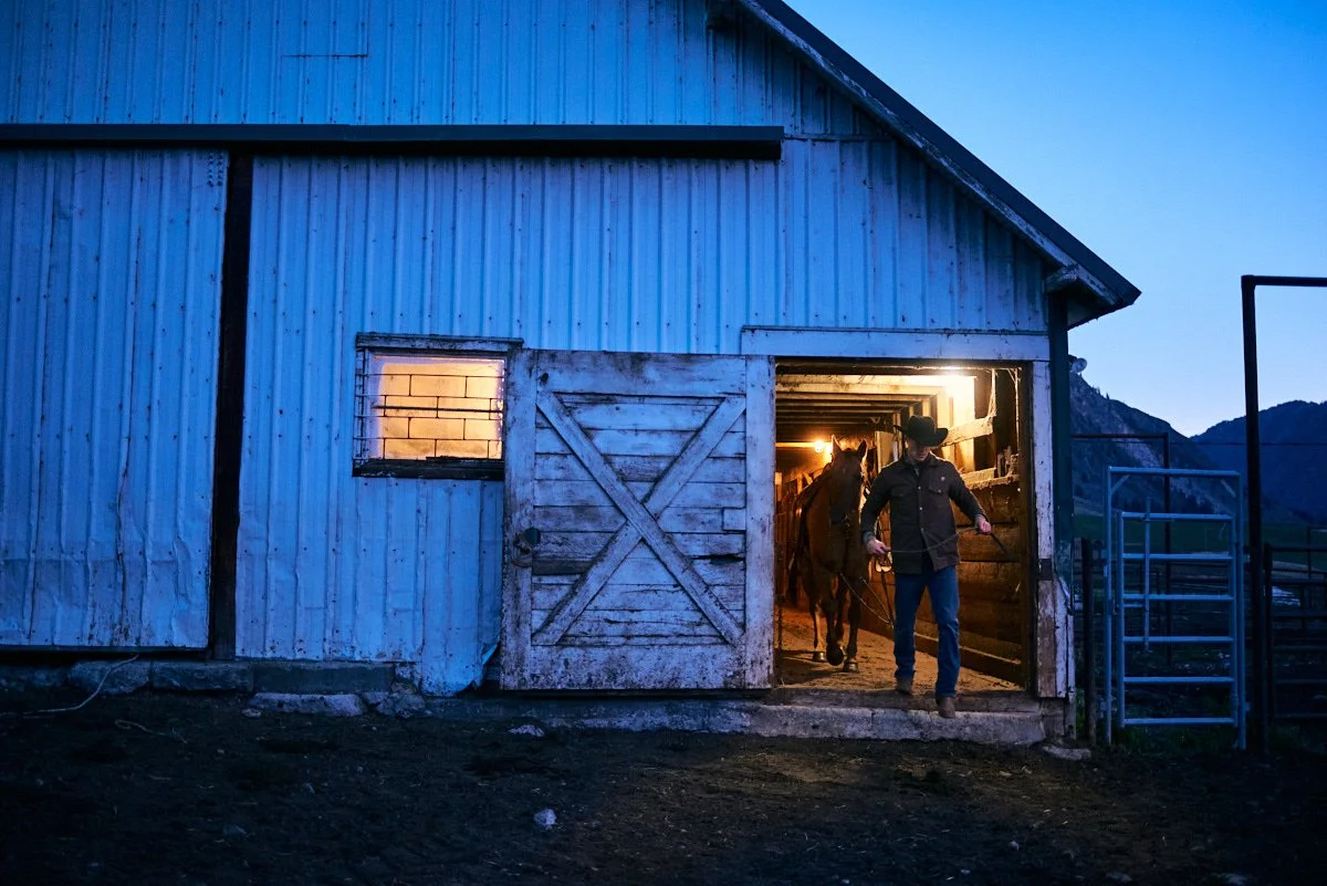 A person in a cowboy hat leading a horse out of a barn at dusk, with mountains in the background.