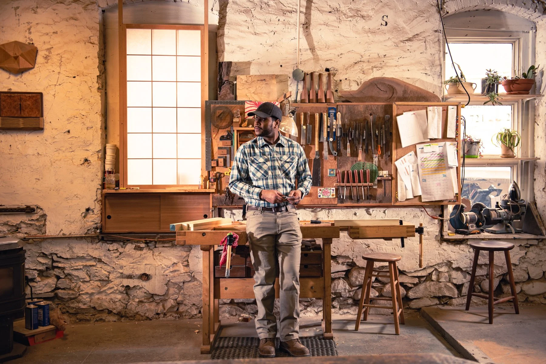 A man standing in a woodworking workshop, surrounded by tools and supplies, with a workbench and windows in the background.