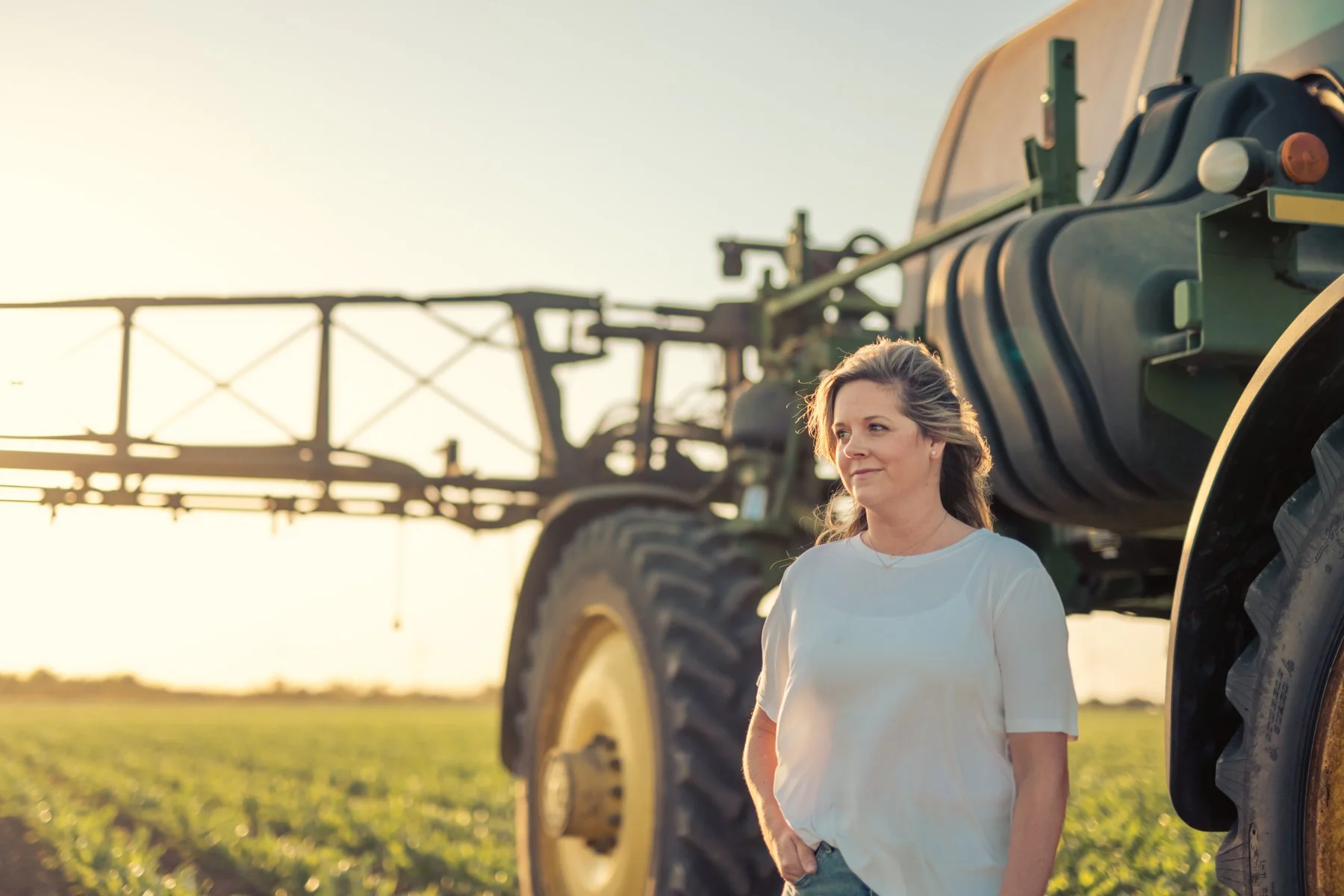 A woman standing in a field with large farming equipment behind her during sunset.