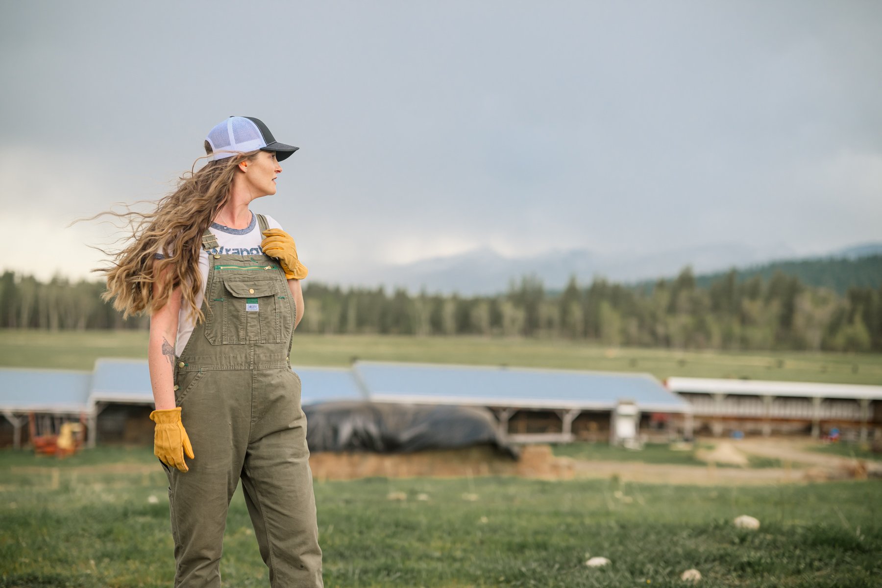 A woman in overalls and a cap stands outdoors with her hand on her shoulder, looking to the side, with a farm and fields in the background under a cloudy sky.