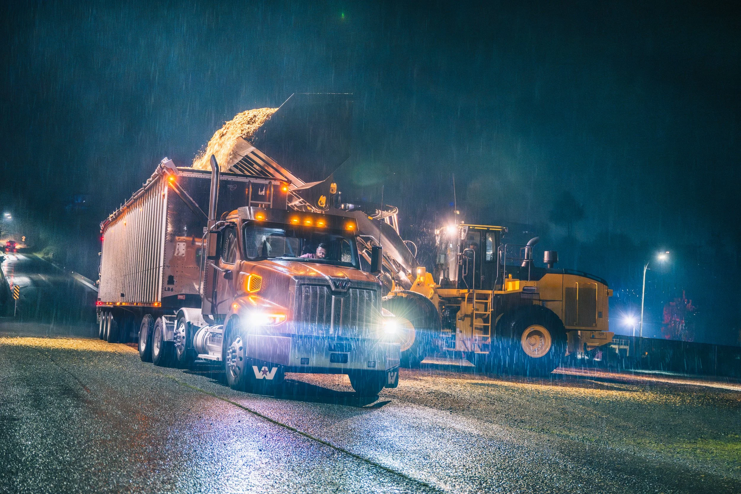 Nighttime scene showing a large dump truck and a road roller working on a wet road, with streetlights and rain visible.