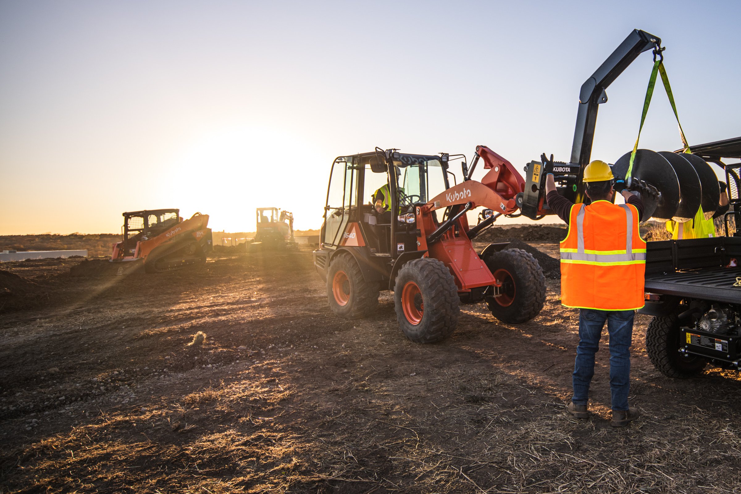 Construction workers operating heavy machinery on a dirt site during sunset.