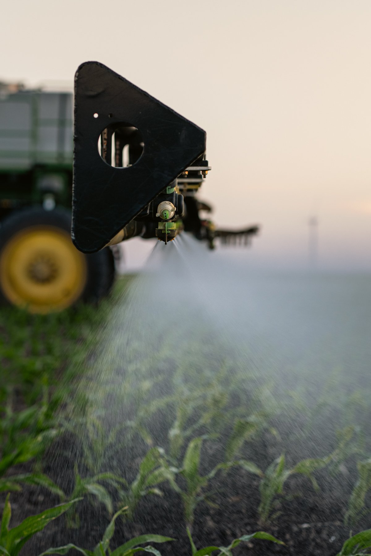 A close-up of a tractor-mounted sprayer applying pesticide or fertilizer on a green crop in a field, with a blurred background of the field and a clear sky.