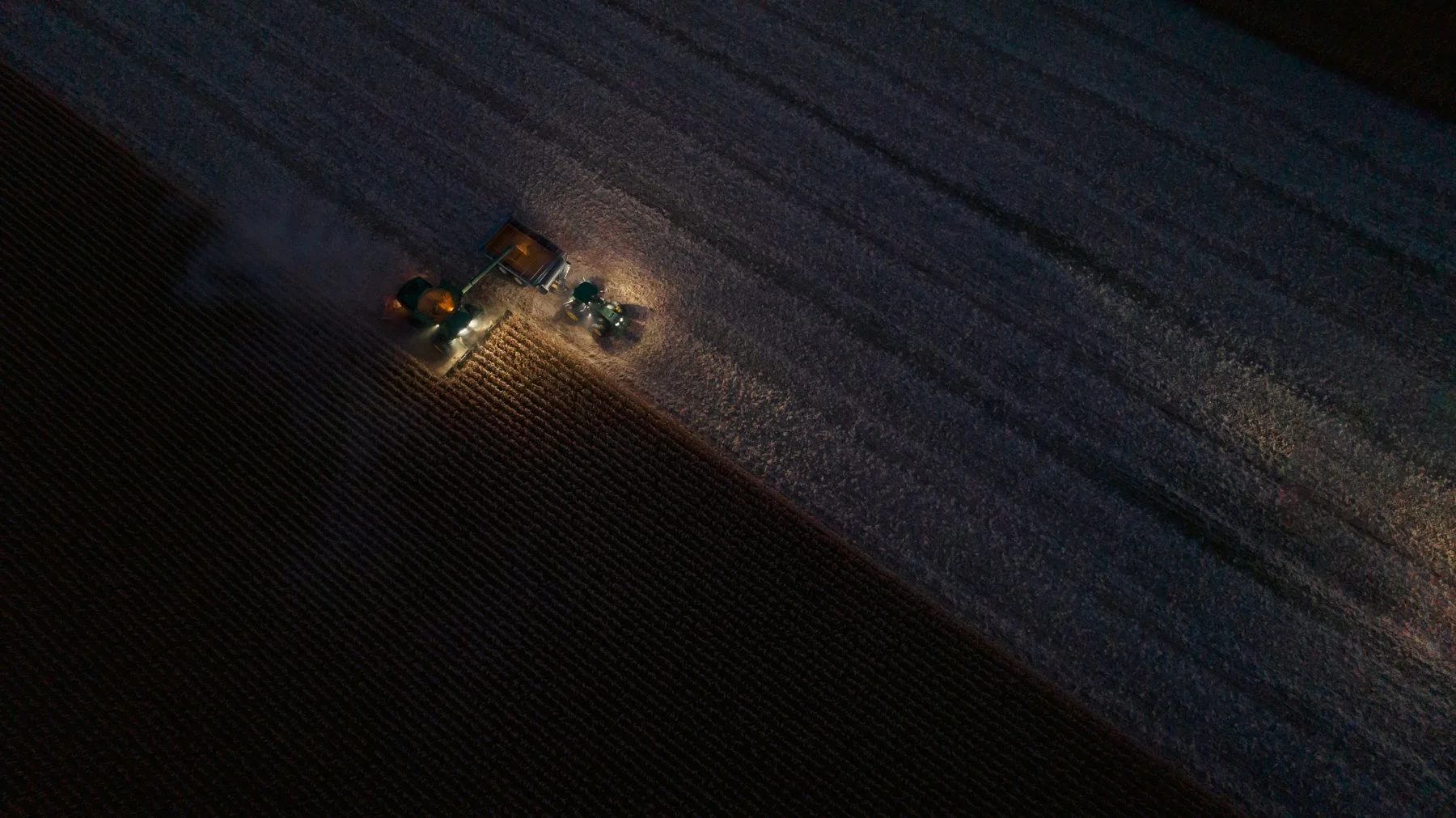 Aerial view of a tractor working in a field at night, illuminated by front lights, with plowed rows visible in the dirt.