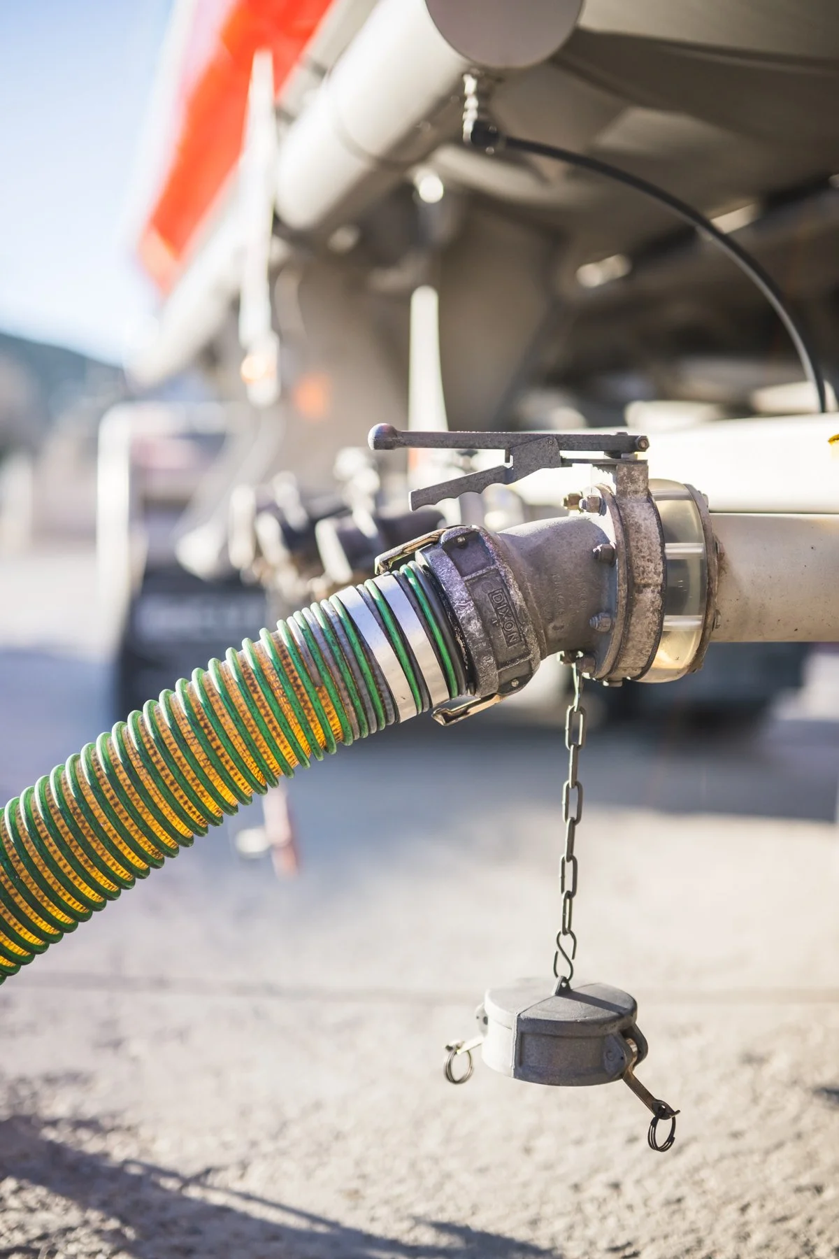 Close-up of a large, flexible hose connected to a metal pipe on a vehicle, with a chain and a small attachment hanging from it, outdoors in bright sunlight.