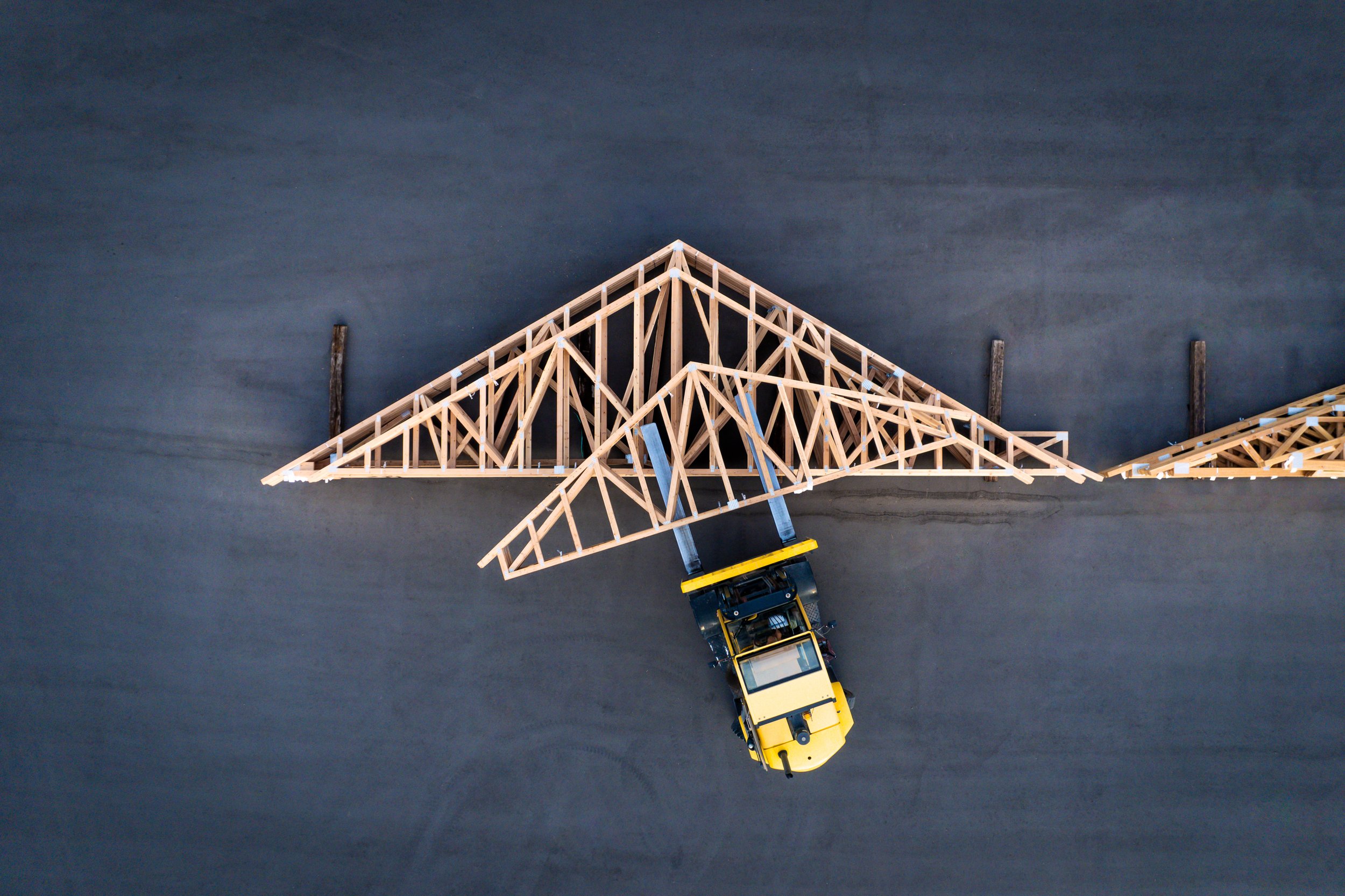 A yellow construction vehicle on a dark asphalt surface lifting a wooden framework for a roof.