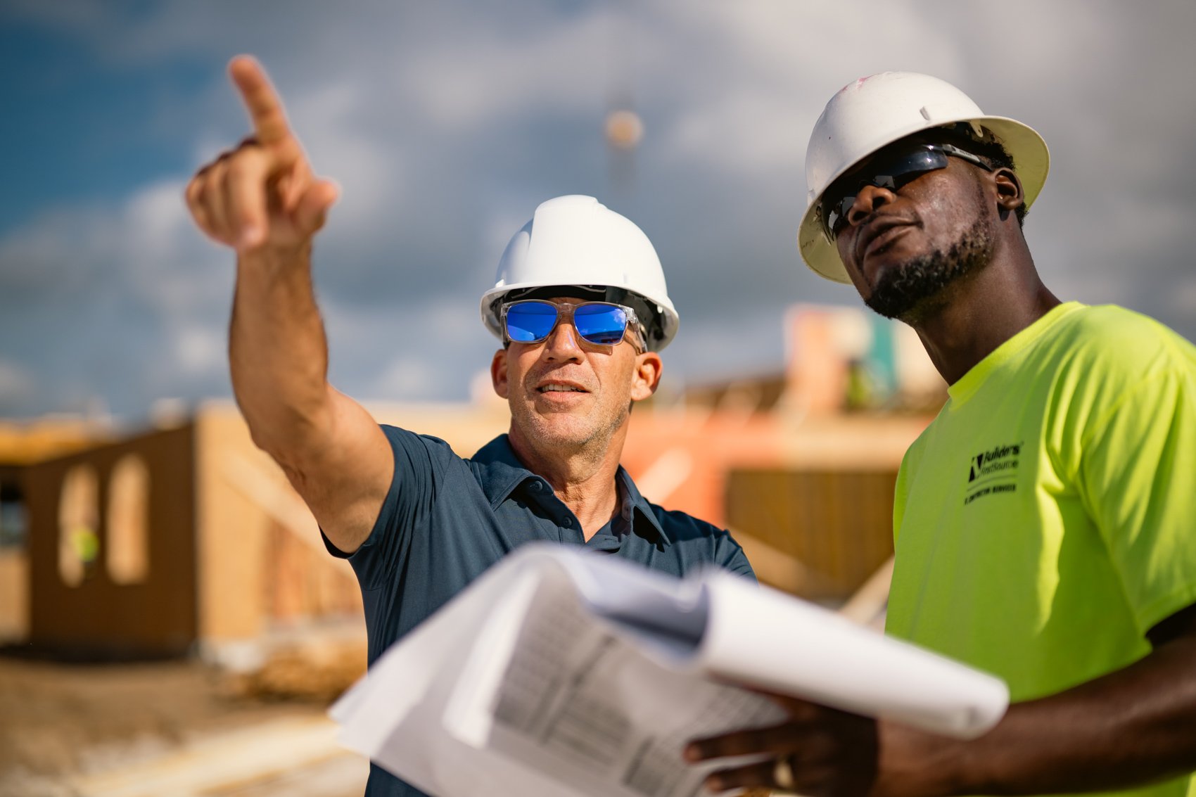 Two construction workers wearing white safety helmets and sunglasses, reviewing blueprints outdoors at a construction site with partially built structures and a cloudy sky in the background.