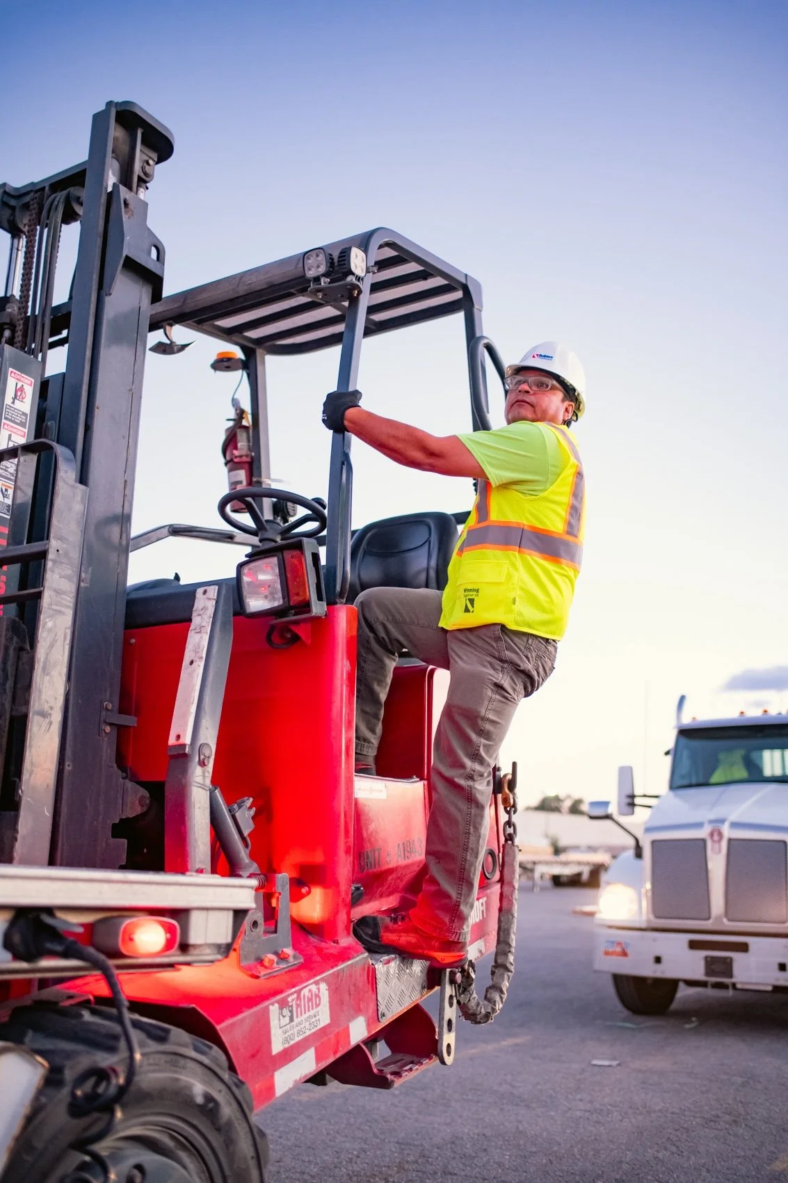 Construction worker wearing safety gear, sitting on a cherry picker lift, with trucks parked in the background at dusk.
