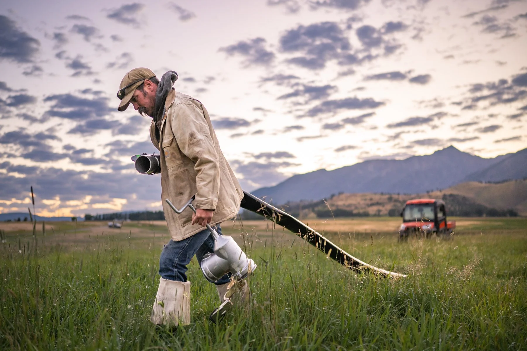 A man dressed in work clothes, carrying a metal watering can, walks through a grassy field with a metal detector hanging around his neck. The setting appears to be rural with mountains in the background and a tractor in the distance, under a cloudy s
