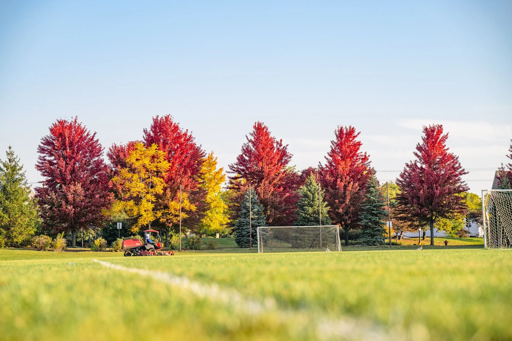 A soccer field in a park with goalposts and a small tractor, surrounded by trees with autumn foliage in red, yellow, and green, under a clear blue sky.