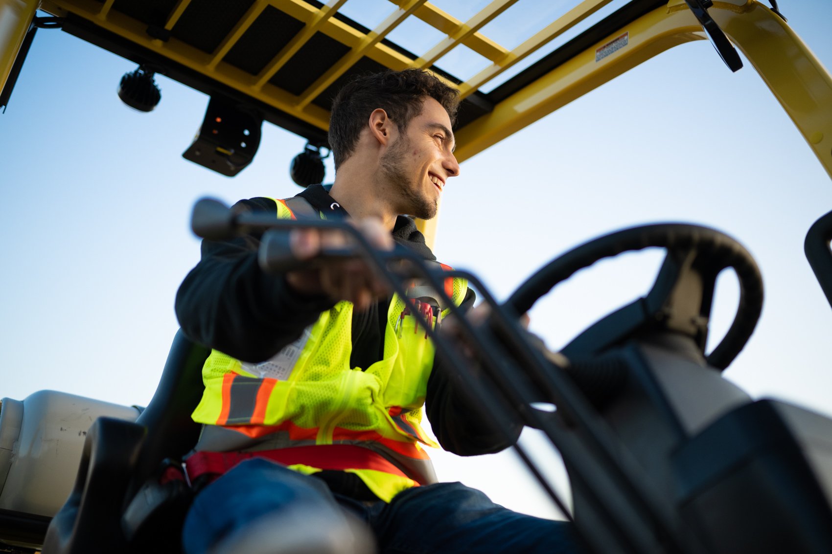 A young man smiling while operating a piece of yellow construction equipment, possibly a skid steer loader, against a clear blue sky.